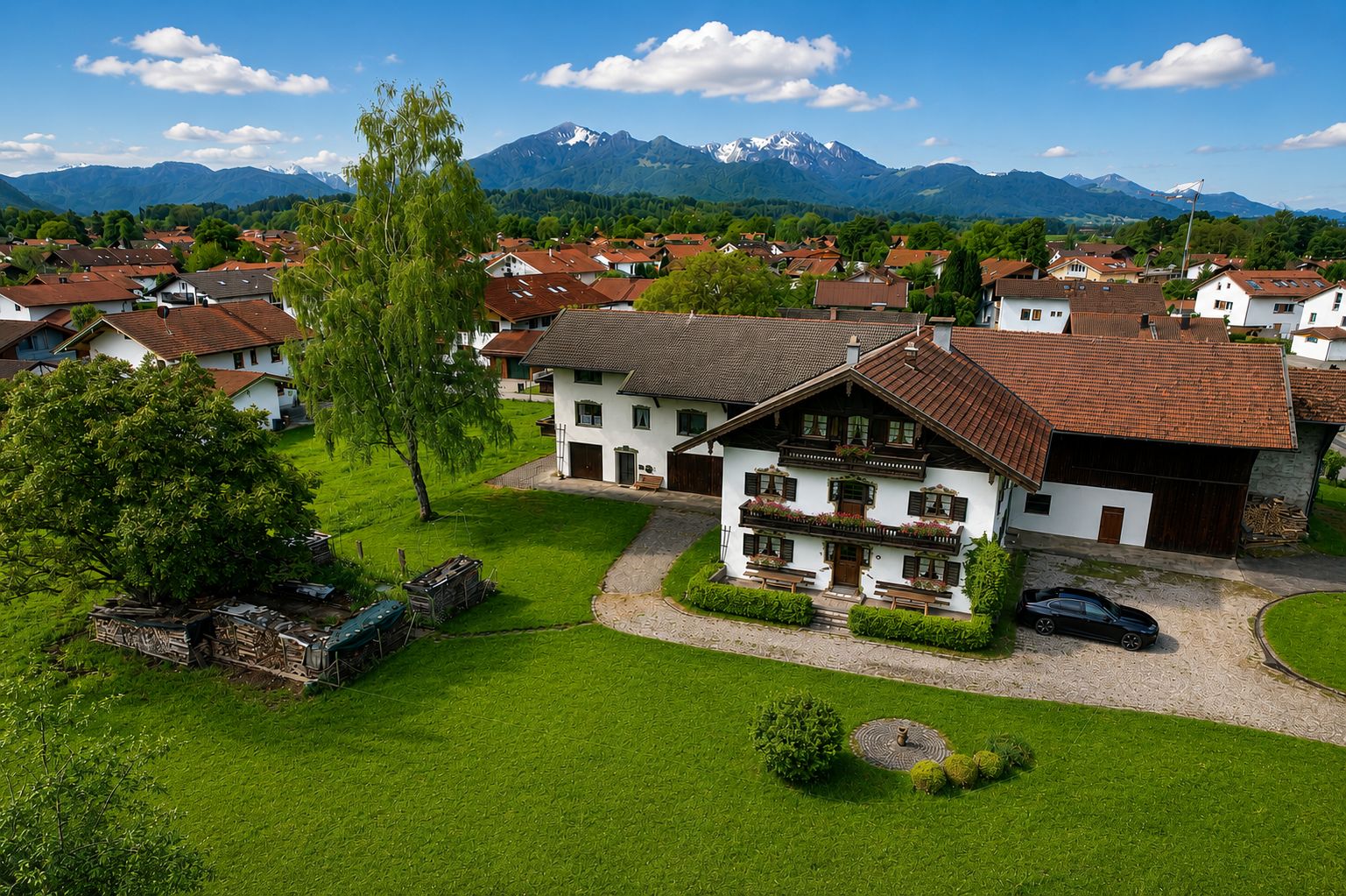 Großes Wohnhaus mit Garten, umgeben von Rasen; im Hintergrund Berge und weitere Häuser.