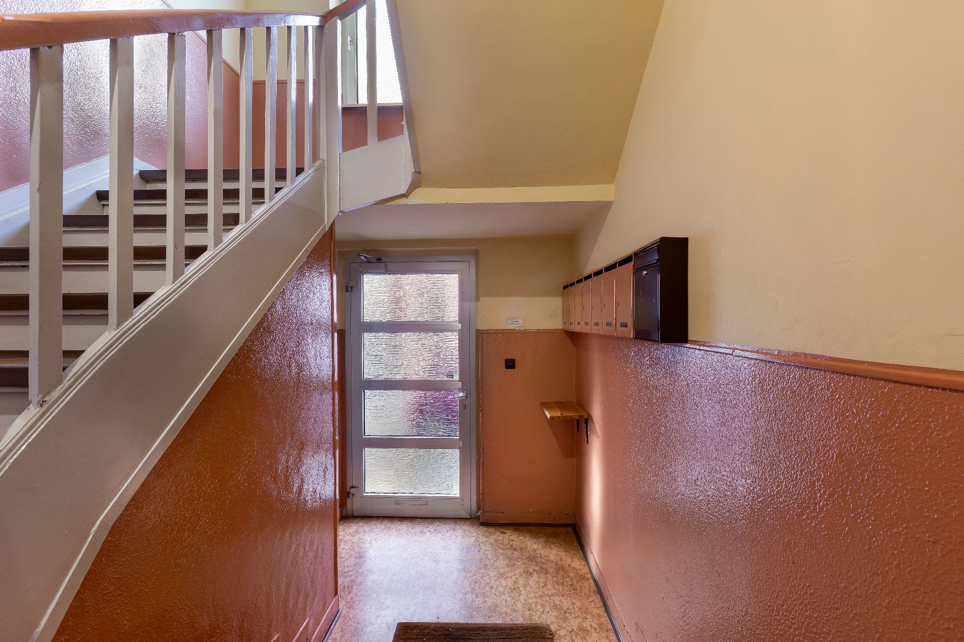 Treppenhaus mit braun-oranger Wand, Glastür und Briefkästen an der Wand.