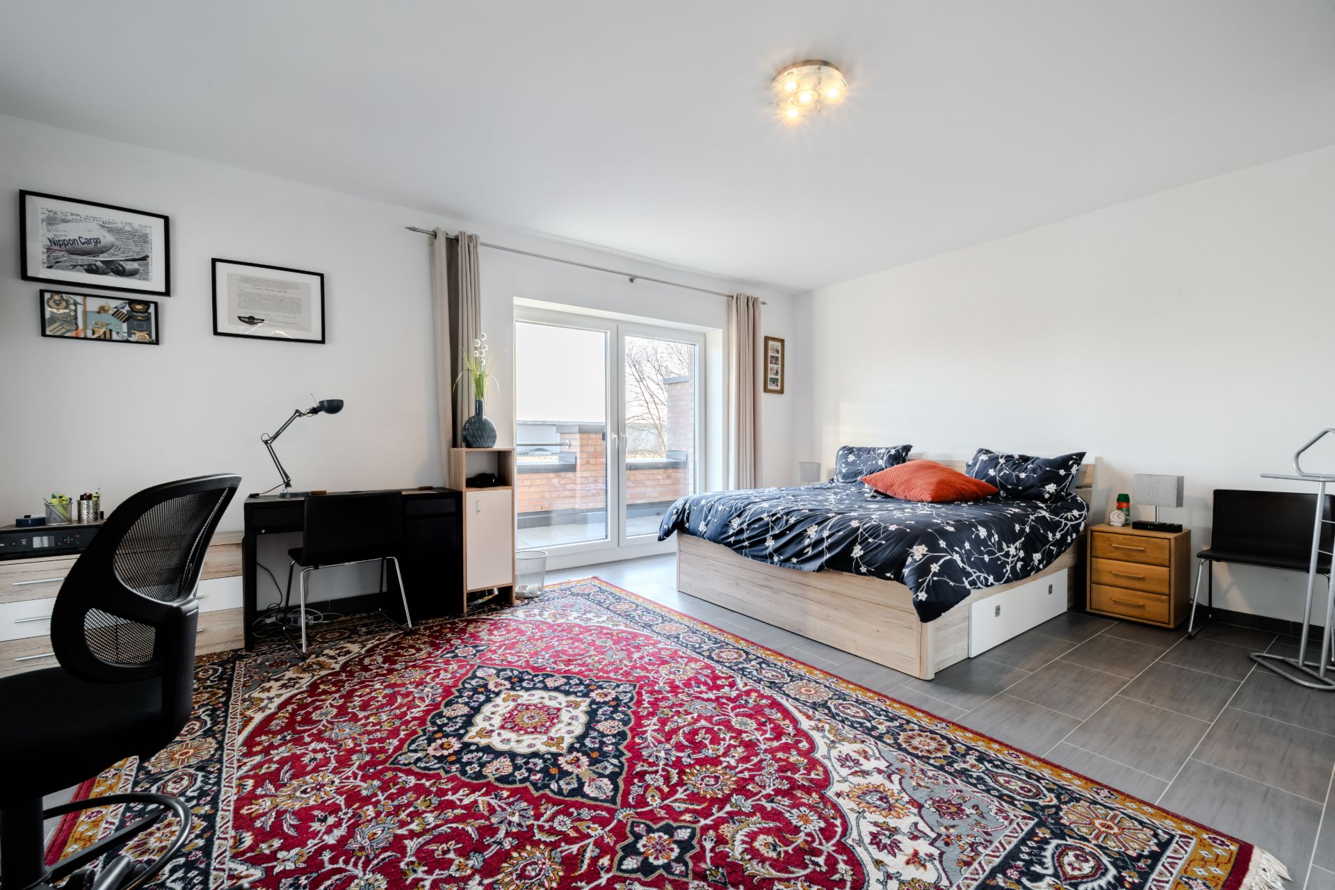 Helles Schlafzimmer mit Doppelbett, Schreibtisch, großem Teppich und Zugang zum Balkon.
