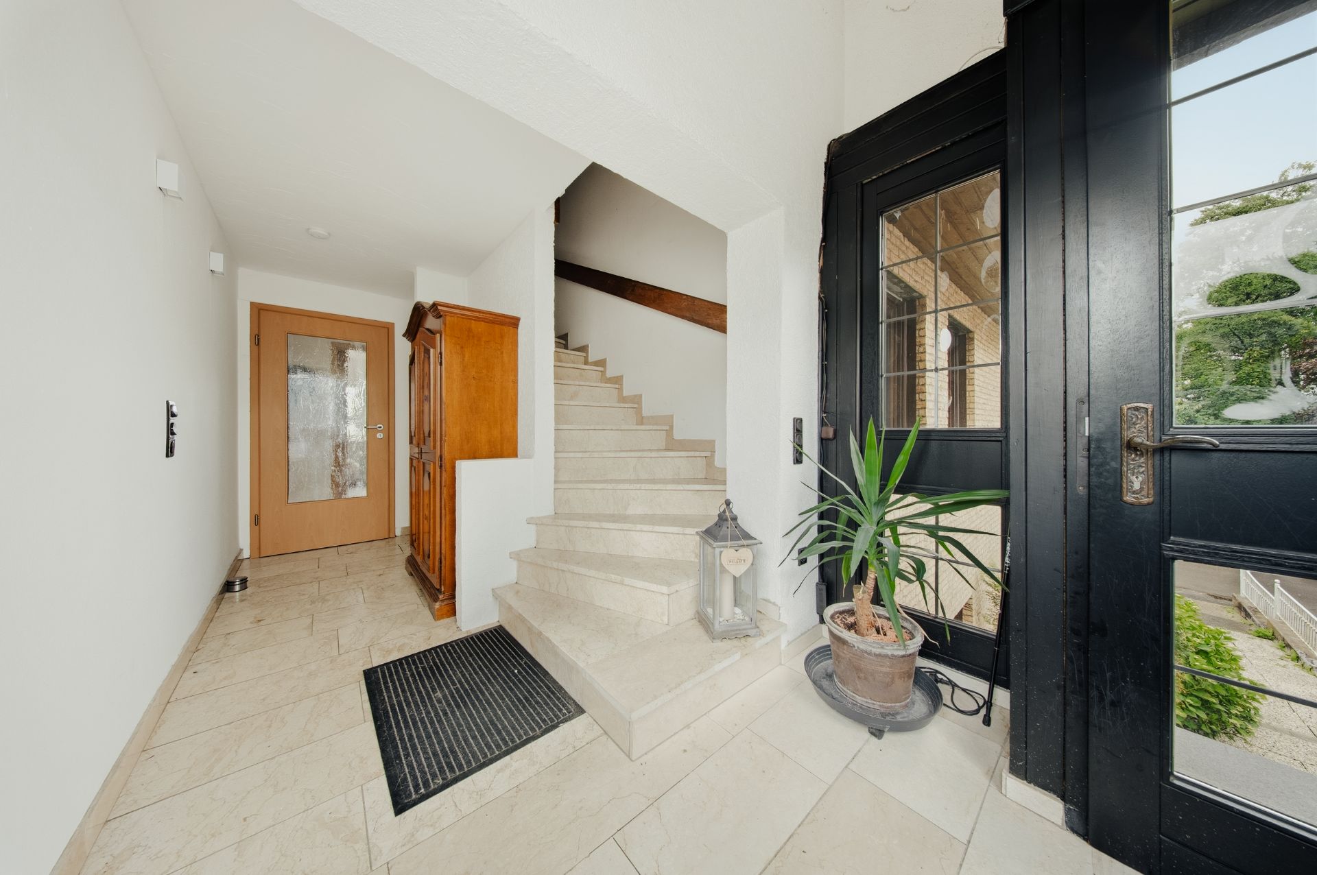 Heller Flur mit Treppe, Holztür, Schrank, Pflanze und großer schwarzer Eingangstür.