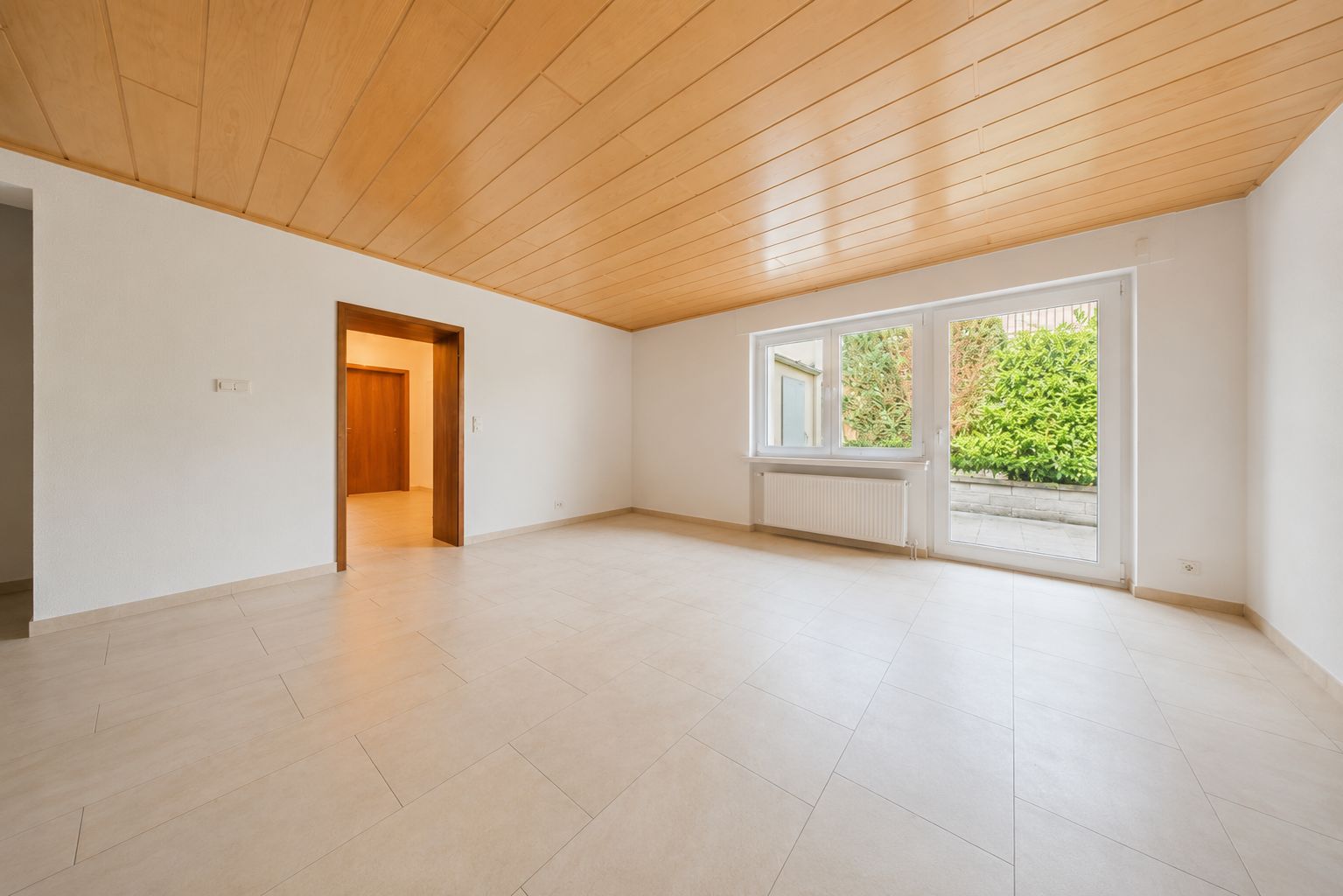 Leeres Wohnzimmer mit heller Fliese, Holzdecke und großem Fenster zum Garten.