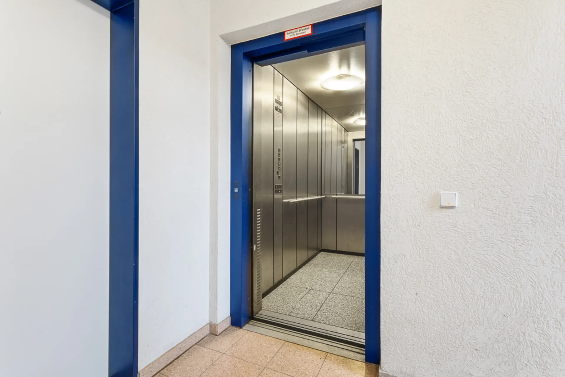 Geöffneter Aufzug mit Edelstahlwänden und blauem Rahmen in einem Wohnhausflur.
