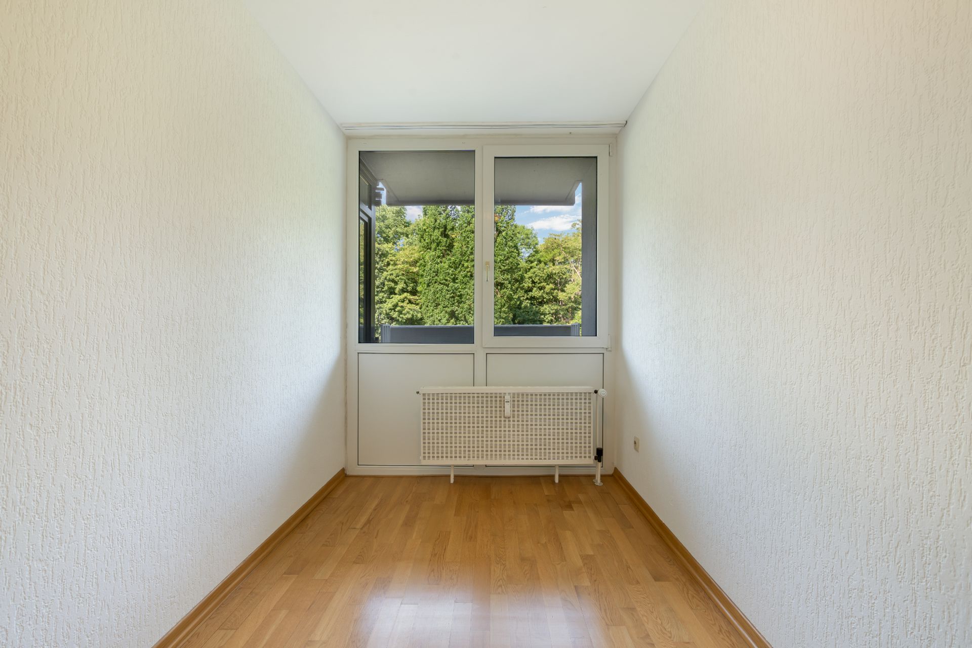 Heller Raum mit Holzboden, weißen Wänden und großem Fenster mit Blick auf Bäume.