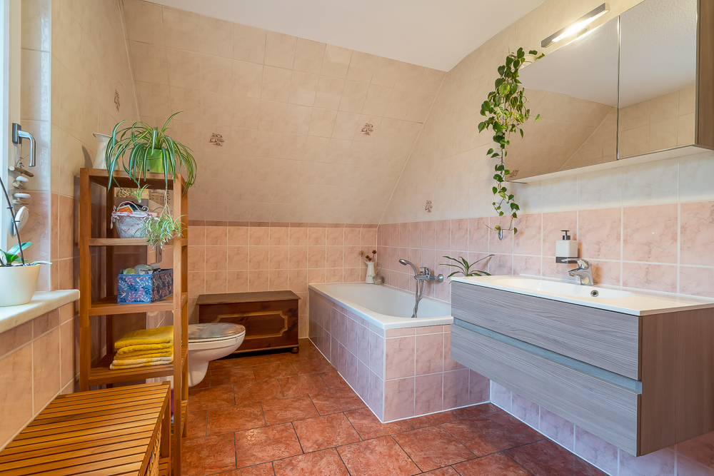 Helles Badezimmer mit Badewanne, Waschbecken, WC, Holzregal und mehreren Zimmerpflanzen.