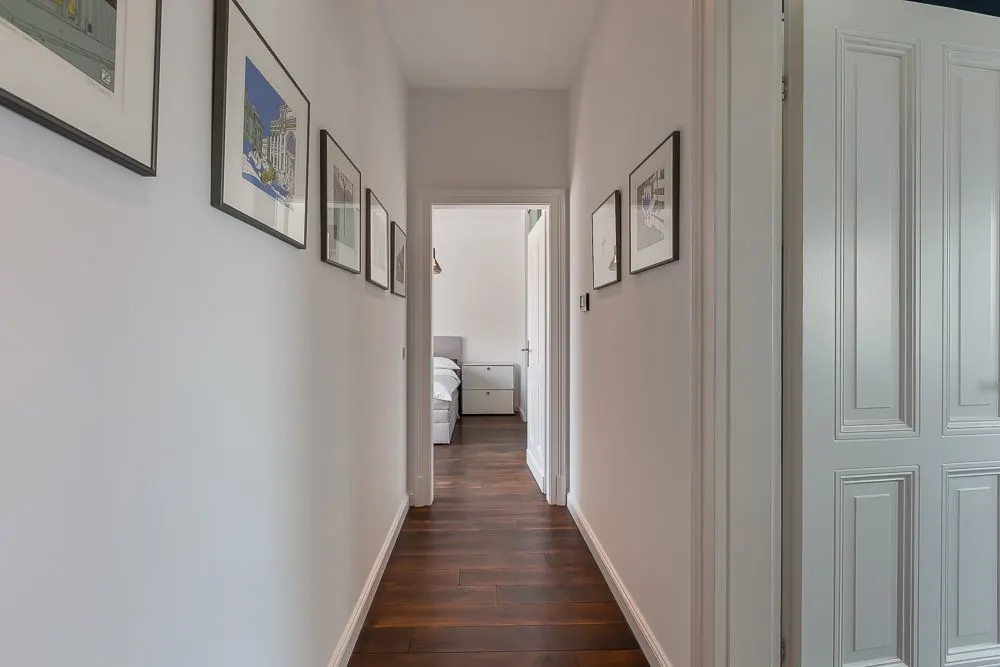 Heller Flur mit Holzboden, weißen Wänden und mehreren Bildern, am Ende ein Schlafzimmer.