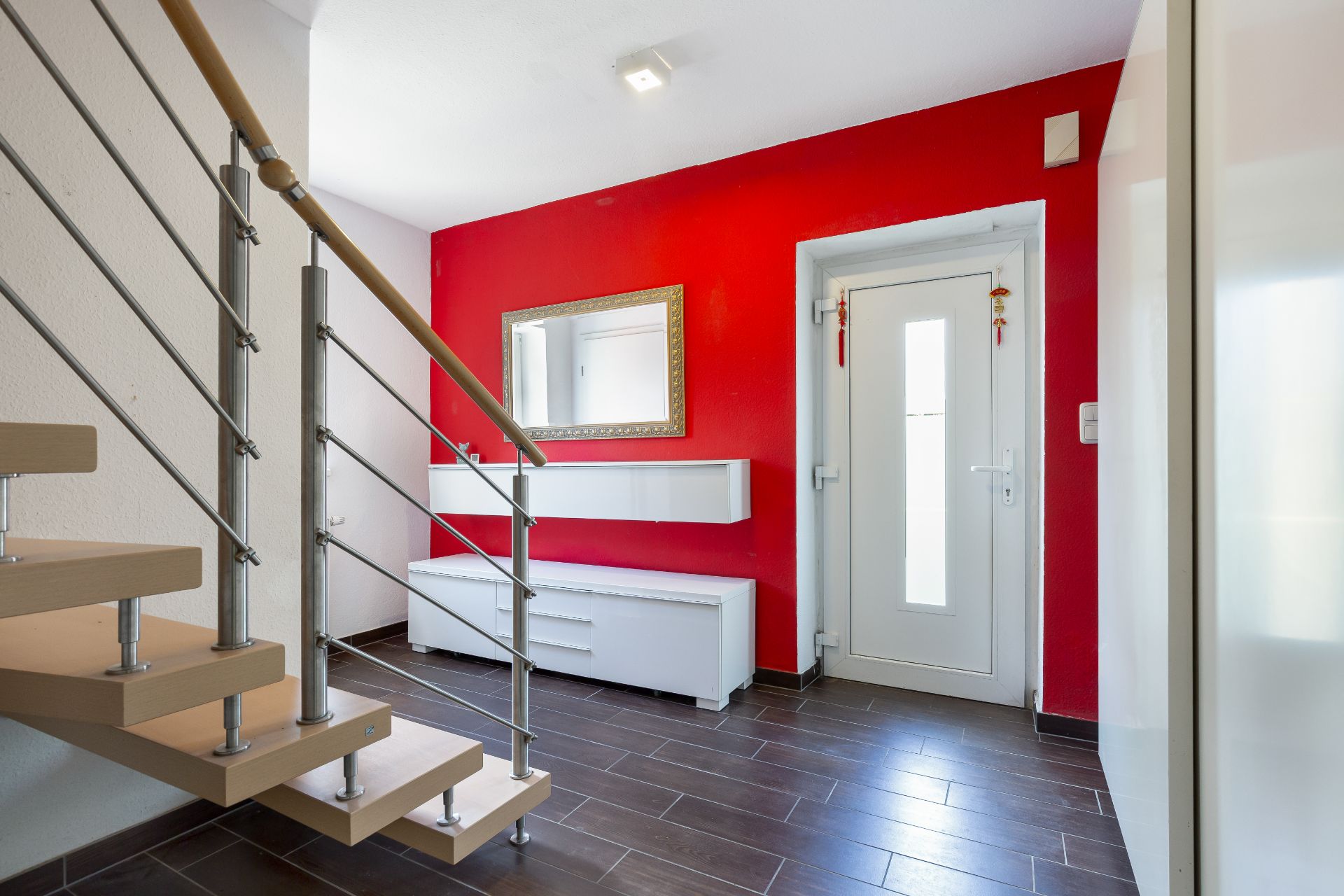 Moderner Flur mit roter Wand, Treppe, Spiegel und weißer Eingangstür.
