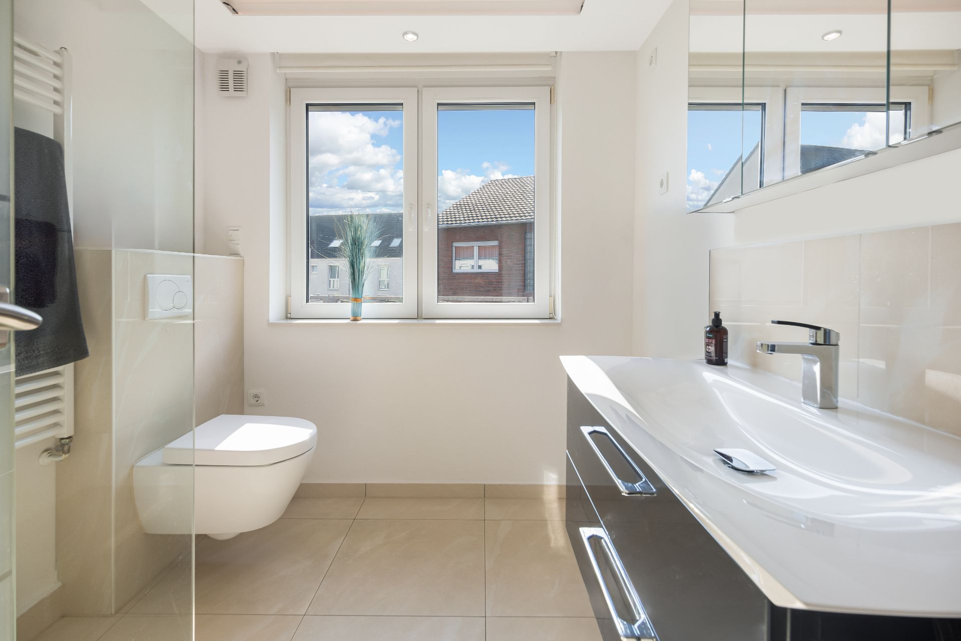 Helles Badezimmer mit Wandtoilette, großem Waschbecken und Fenster mit Blick nach draußen.