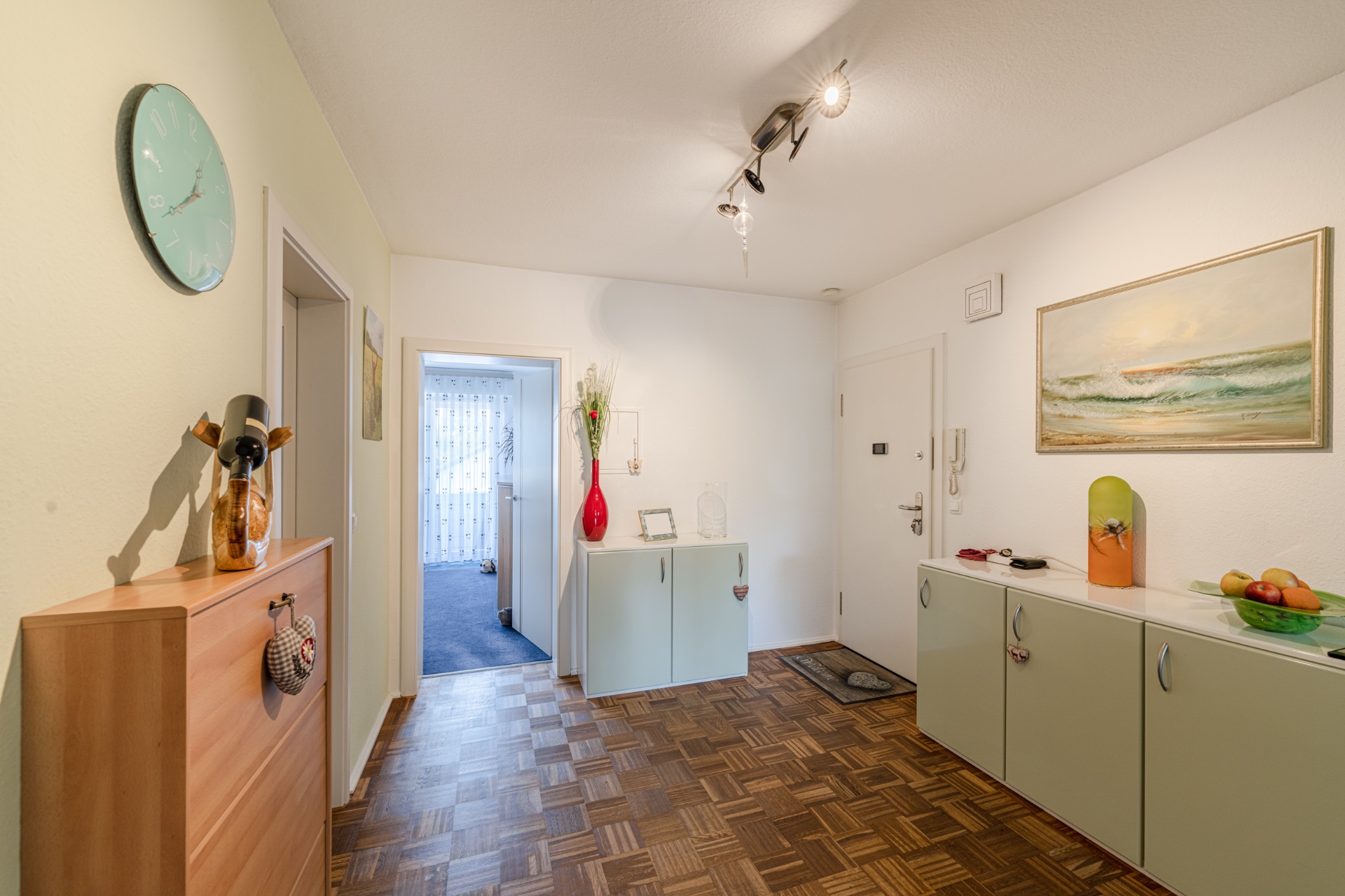 Heller Flur mit Holzparkett, Sideboards, Wandbild, Uhr und roten Dekovasen.
