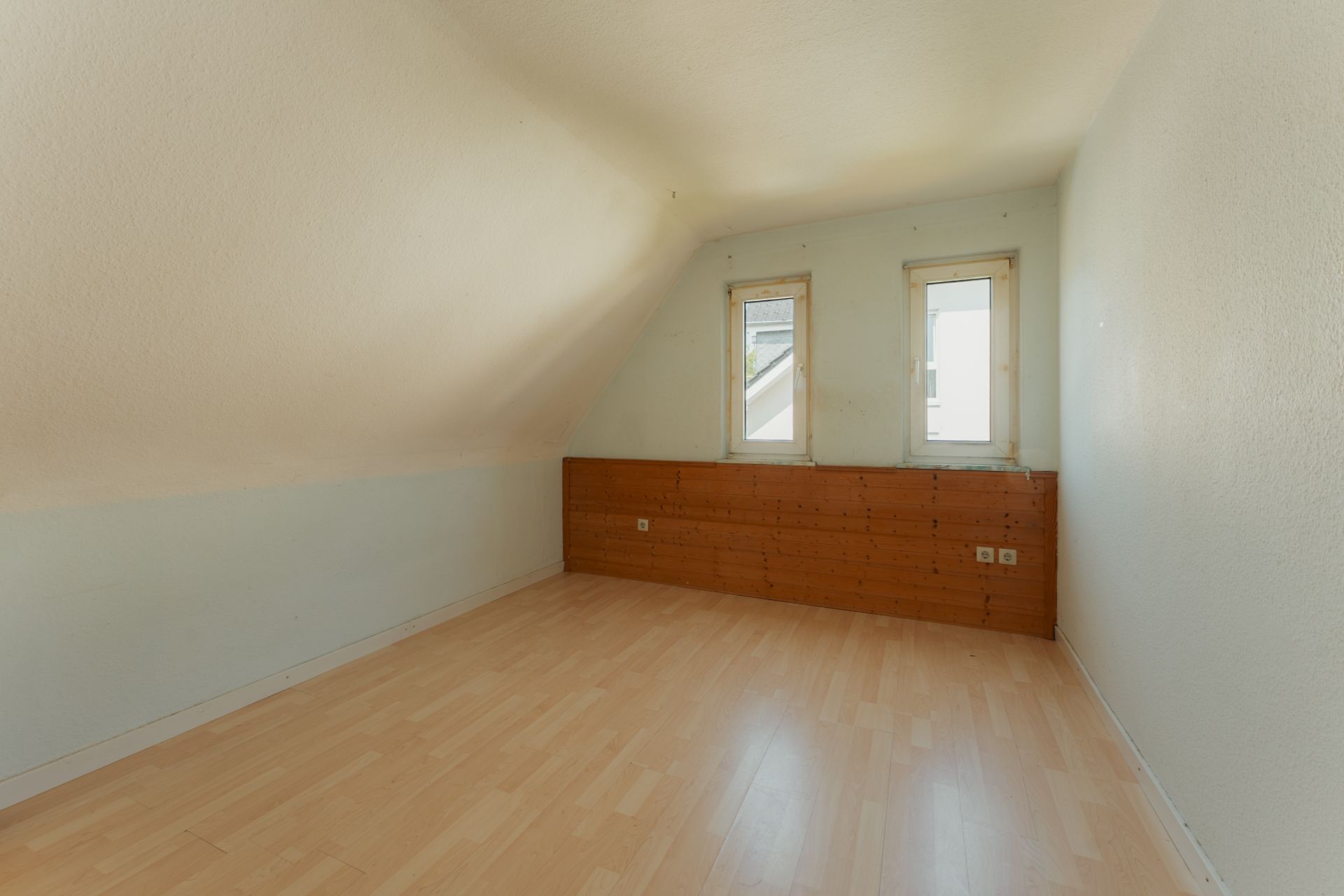Leeres Zimmer mit schräger Decke, zwei Fenstern und hellem Holzboden.