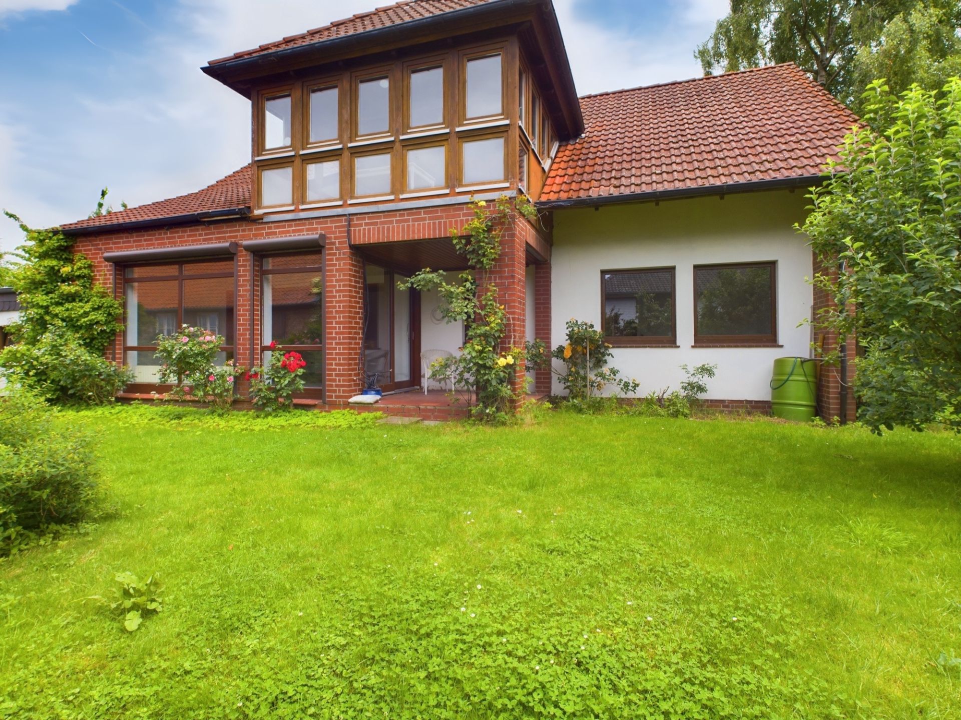 Haus mit rotem Ziegeldach, großem Garten und vielen Fenstern, etwas Blumen im Vorgarten.