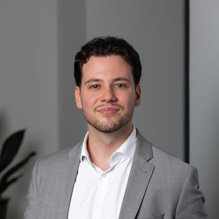 Ein Mann in grauem Anzug und weißem Hemd steht vor einem neutralen Hintergrund.