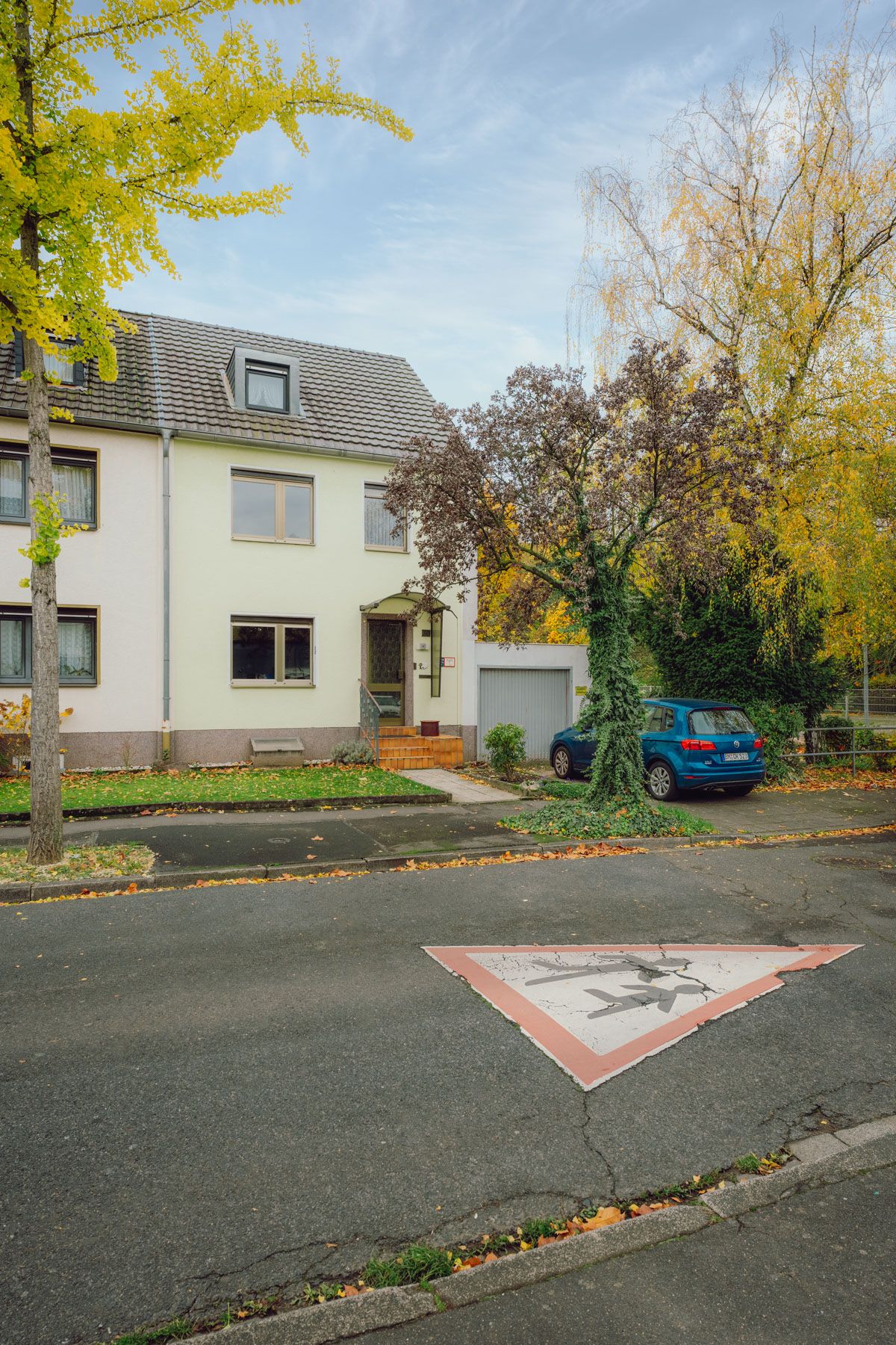 Wohnhaus mit Garage, davor steht ein blaues Auto, Bäume mit Herbstlaub und Zebrastreifen-Straßenmarkierung.