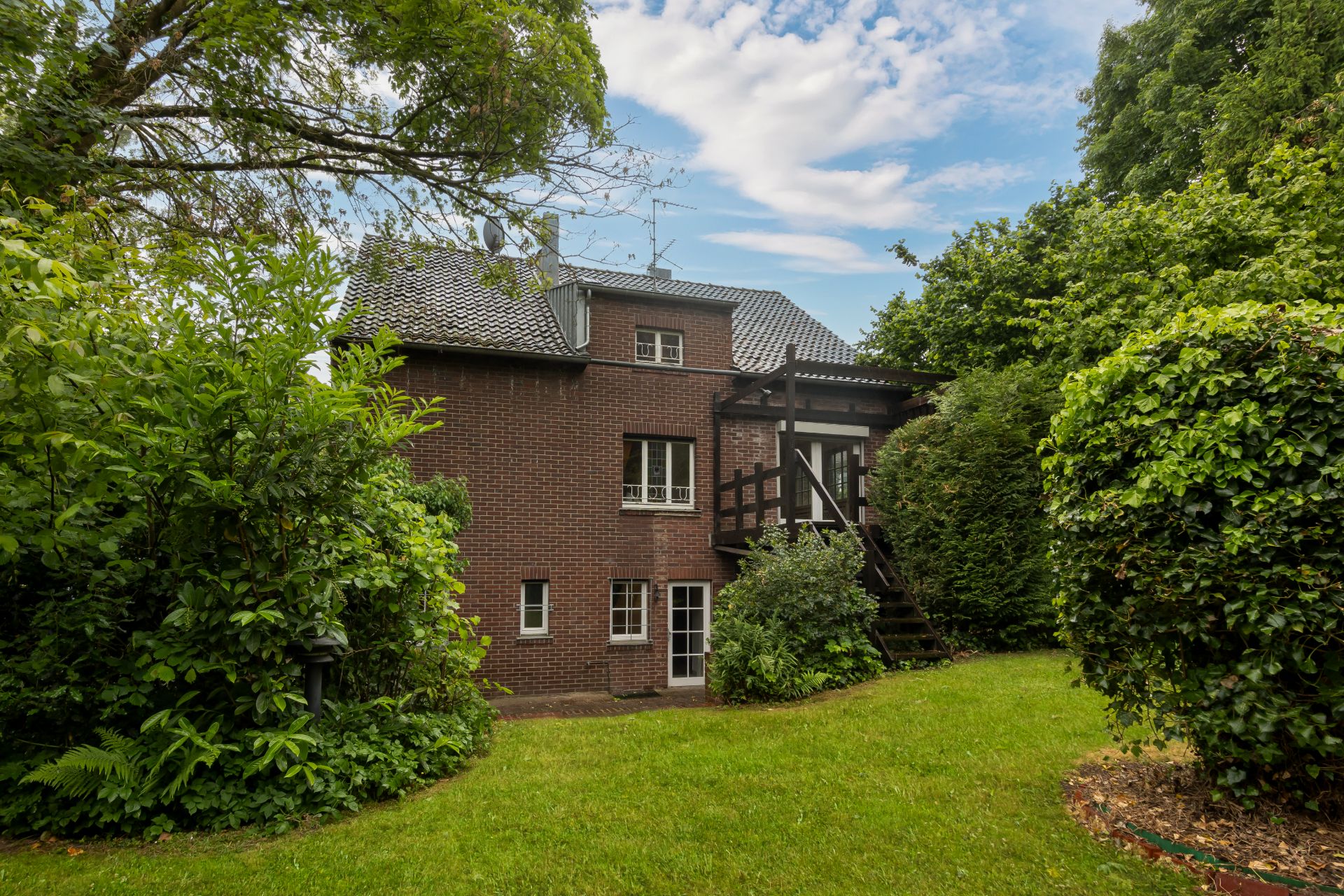 Backsteinwohnhaus mit Balkon und Treppe, umgeben von viel Grün und Bäumen.