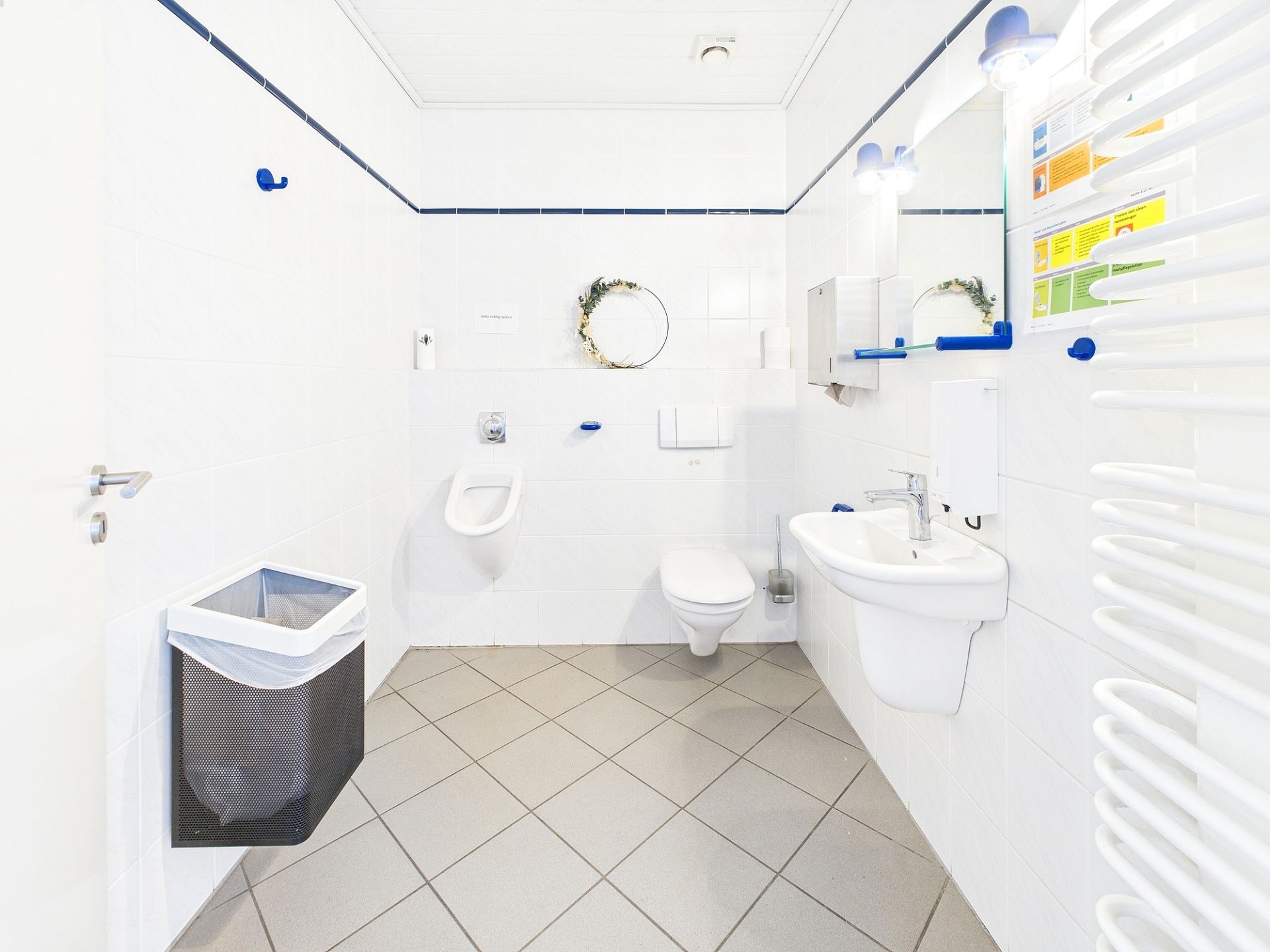 Helles Badezimmer mit Waschbecken, Toilette, Urinal, Spiegel und Mülleimer.