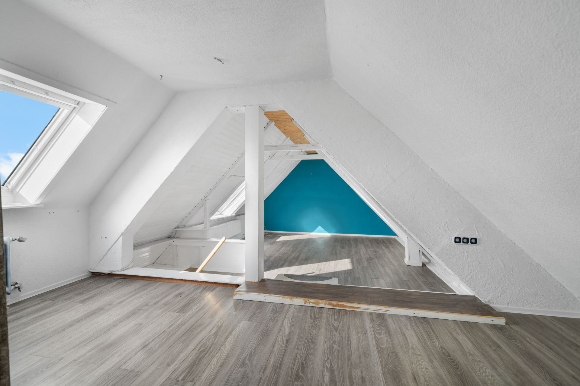 Heller Dachgeschossraum mit Fenster, schrägen Wänden und blau gestrichener Wand im Hintergrund.