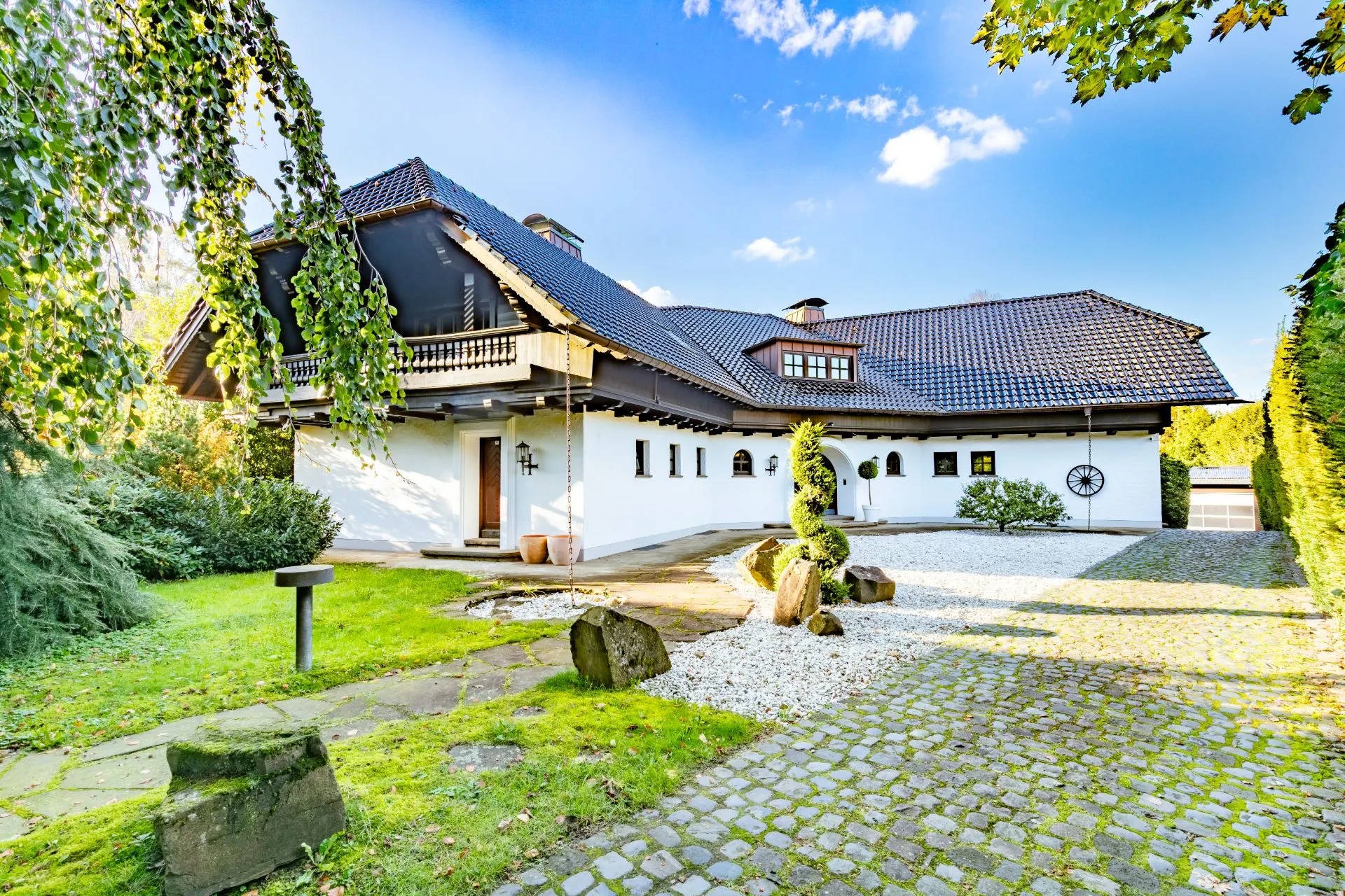 Großes weißes Wohnhaus mit dunklem Dach, Garten, Kiesweg und Kopfsteinpflaster im Vordergrund.