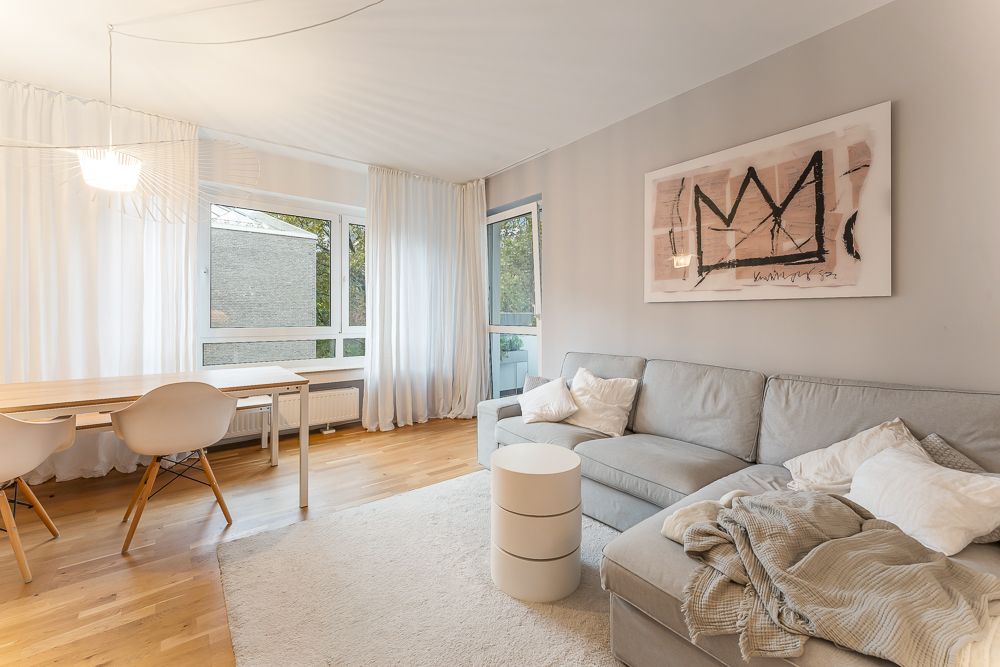 Helles Wohnzimmer mit grauem Sofa, Esstisch, zwei Stühlen und großem Fenster mit weißen Vorhängen.