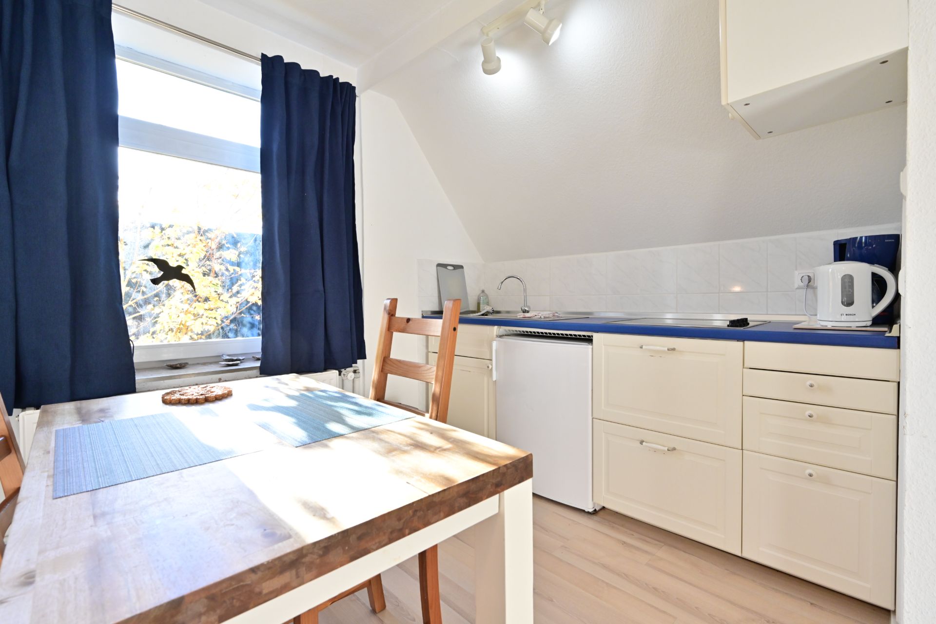 Kleine Küche mit Esstisch, zwei Stühlen, Kühlschrank und Fenster mit blauen Vorhängen.