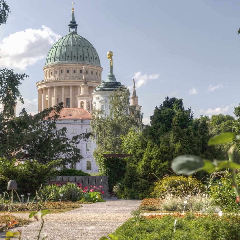 Gepflegter Garten mit Blumen und Sträuchern, im Hintergrund ein großes Gebäude mit Kuppel.