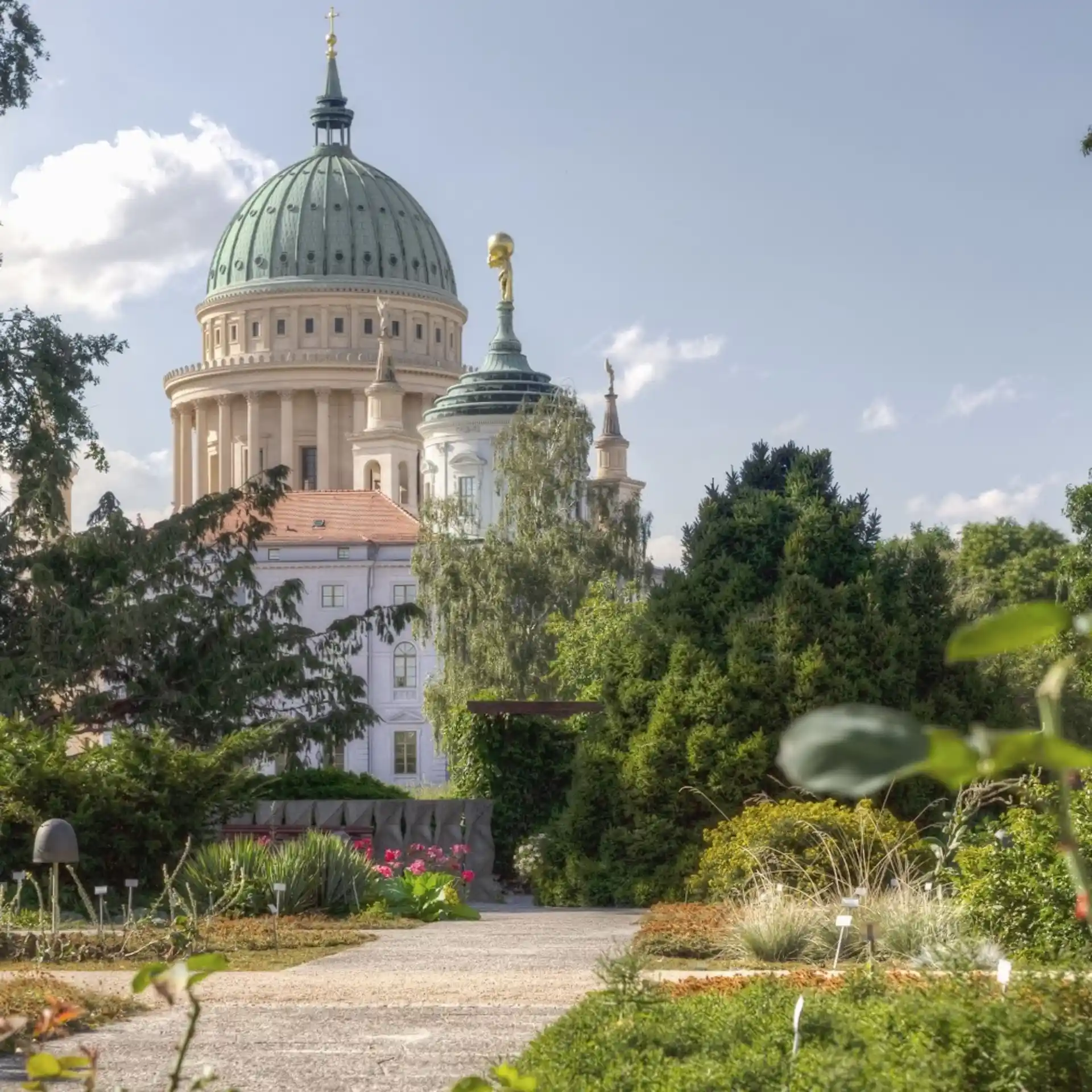 Gepflegter Garten mit Blumen und Sträuchern, im Hintergrund ein großes Gebäude mit Kuppel.
