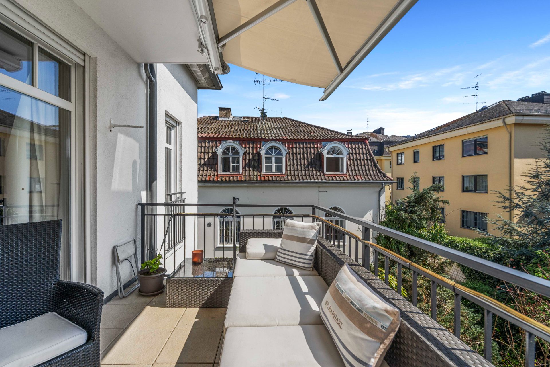Balkon mit Sofa, Stühlen, Pflanzen und Markise, Blick auf Nachbarhäuser bei Tageslicht.