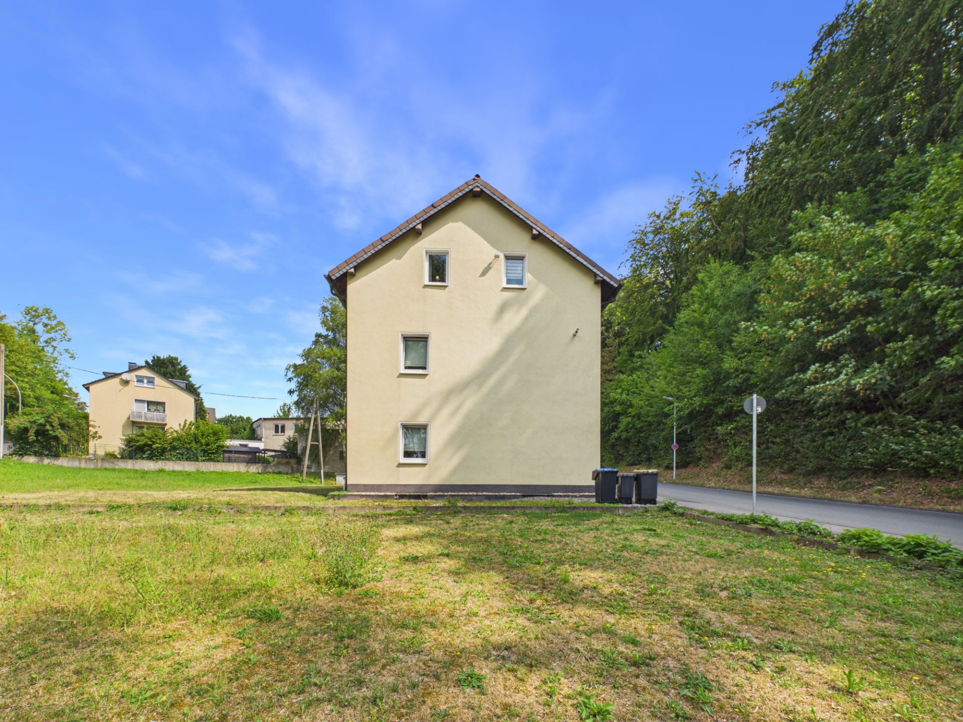 Beiges Wohnhaus mit drei Fenstern, davor Wiese und Straße, umgeben von Bäumen.