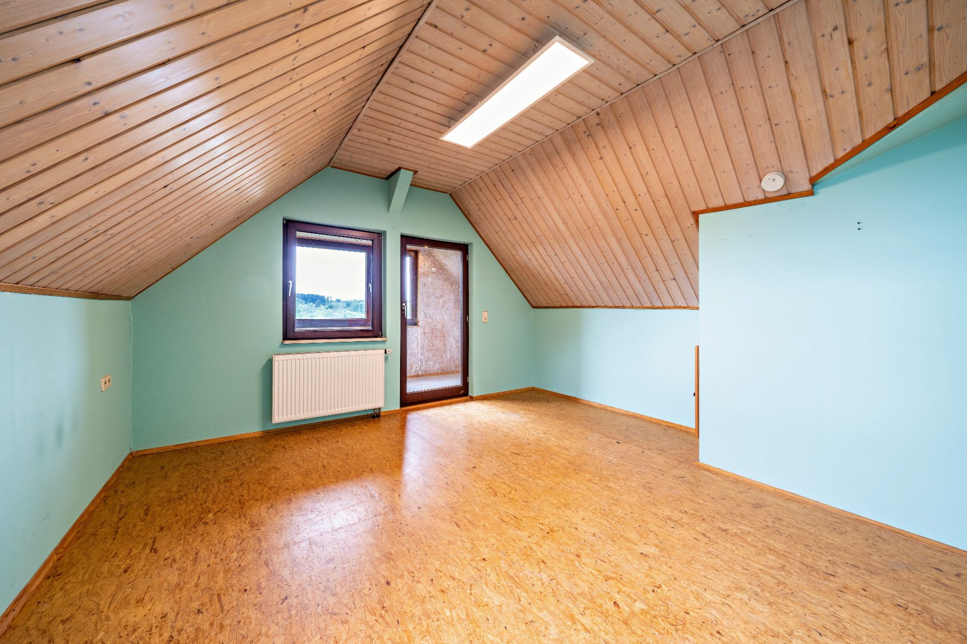 Leerer Dachgeschossraum mit Holzdecke, Fenster, Tür und hellblauen Wänden.