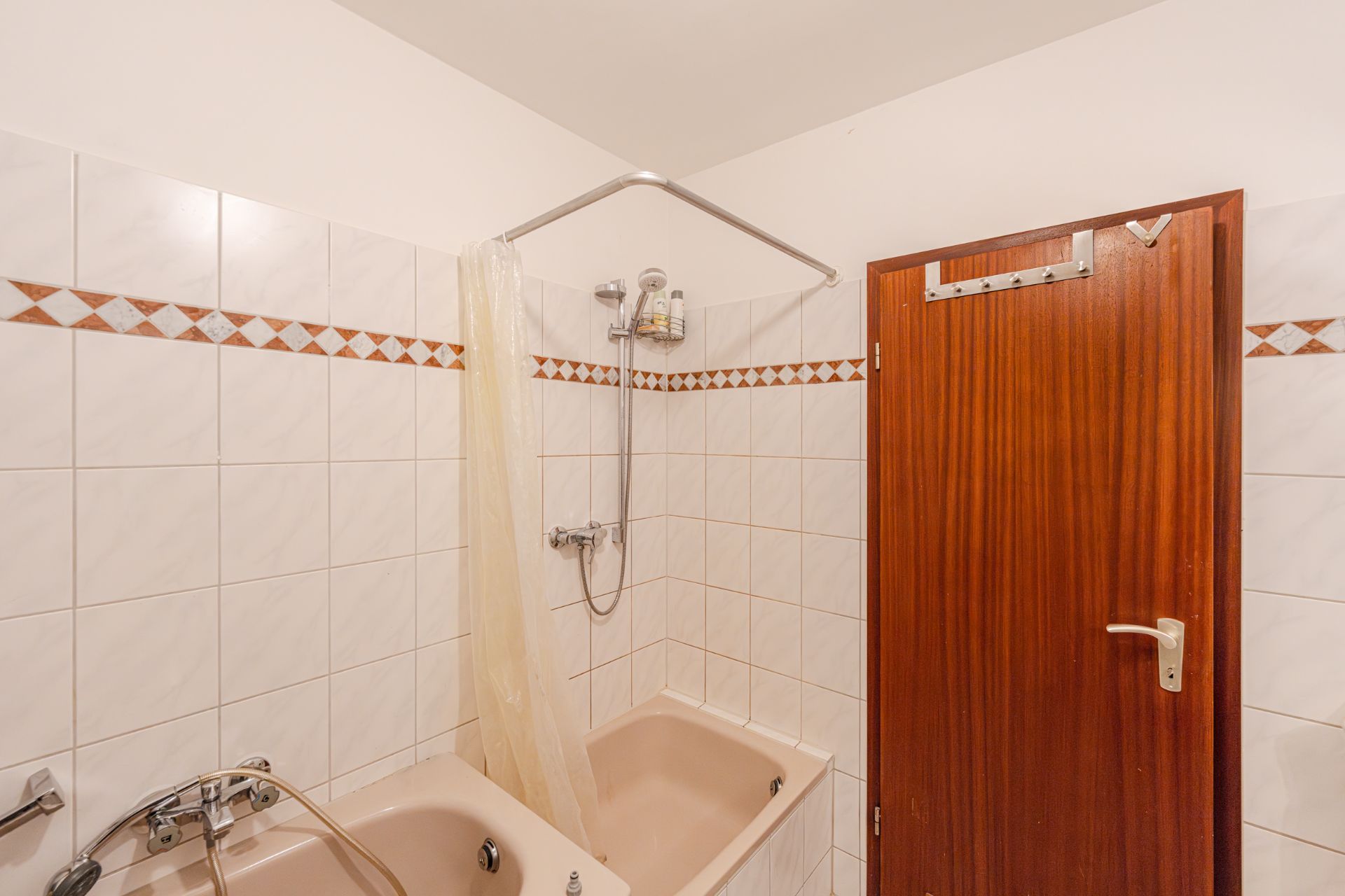 Badezimmer mit Badewanne, Dusche, Duschvorhang und Holztür, weiße Fliesen mit Musterleiste.