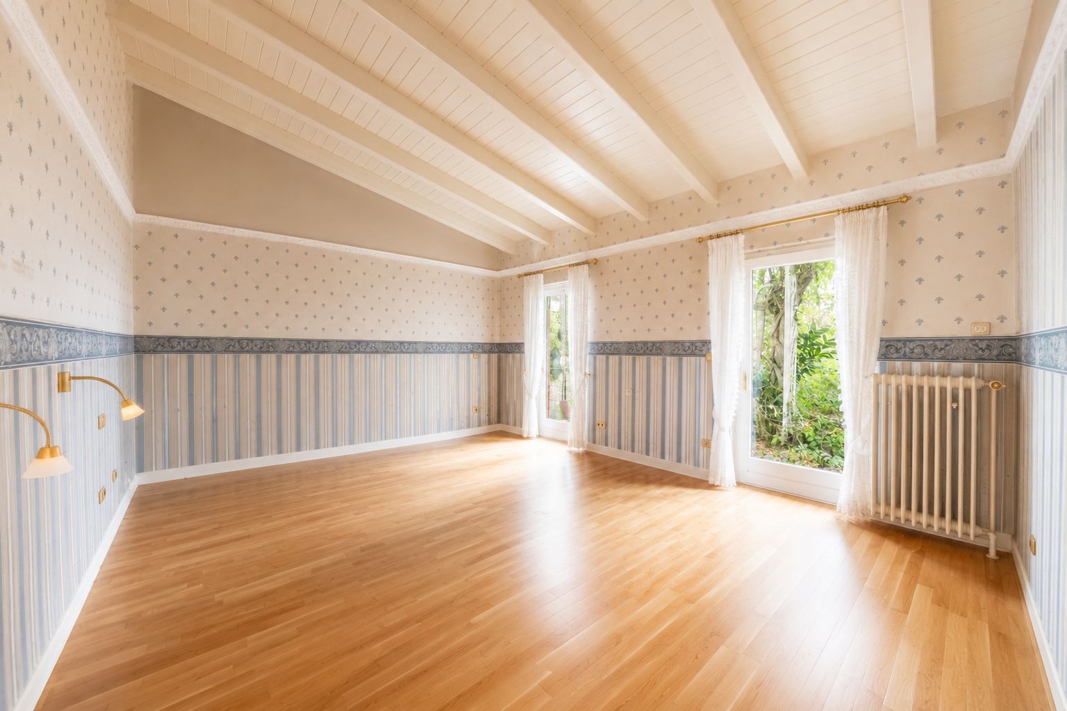 Heller, leerer Raum mit Holzfußboden, Tapeten, großen Fenstern und sichtbaren Deckenbalken.