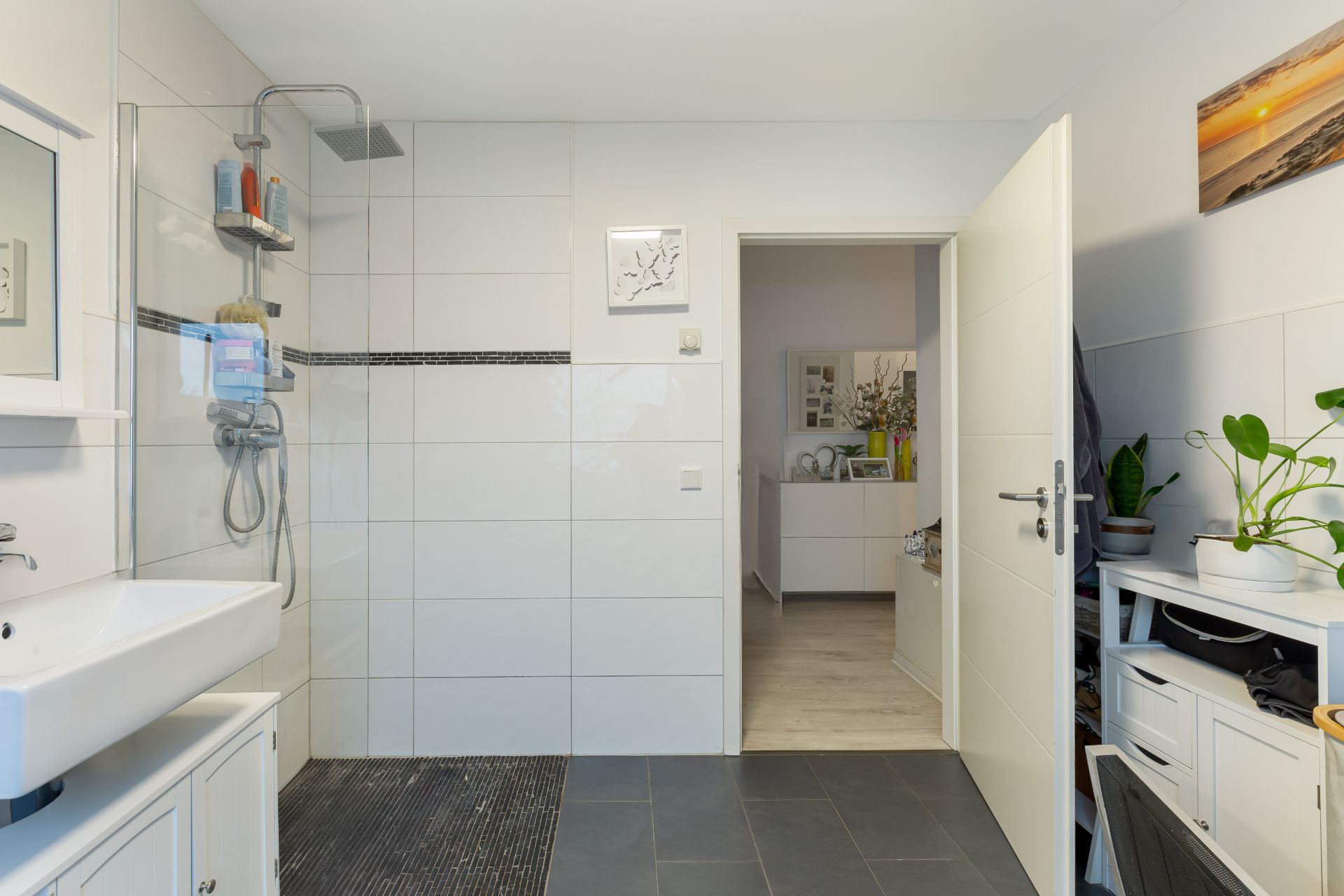 Helles Badezimmer mit ebenerdiger Dusche, Waschbecken und Blick durch offene Tür in einen Flur.