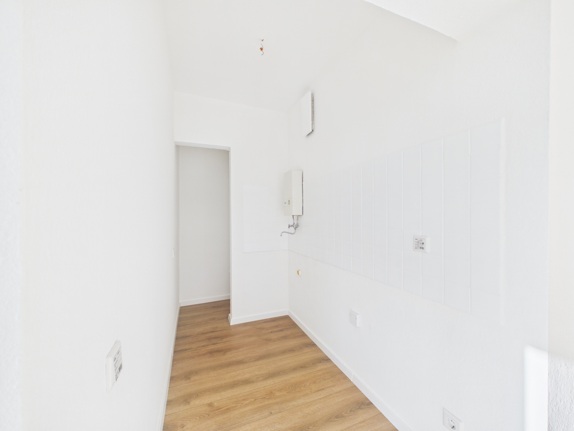 Schmaler, leerer Raum mit weißer Wand und Holzboden, Türöffnung am Ende.
