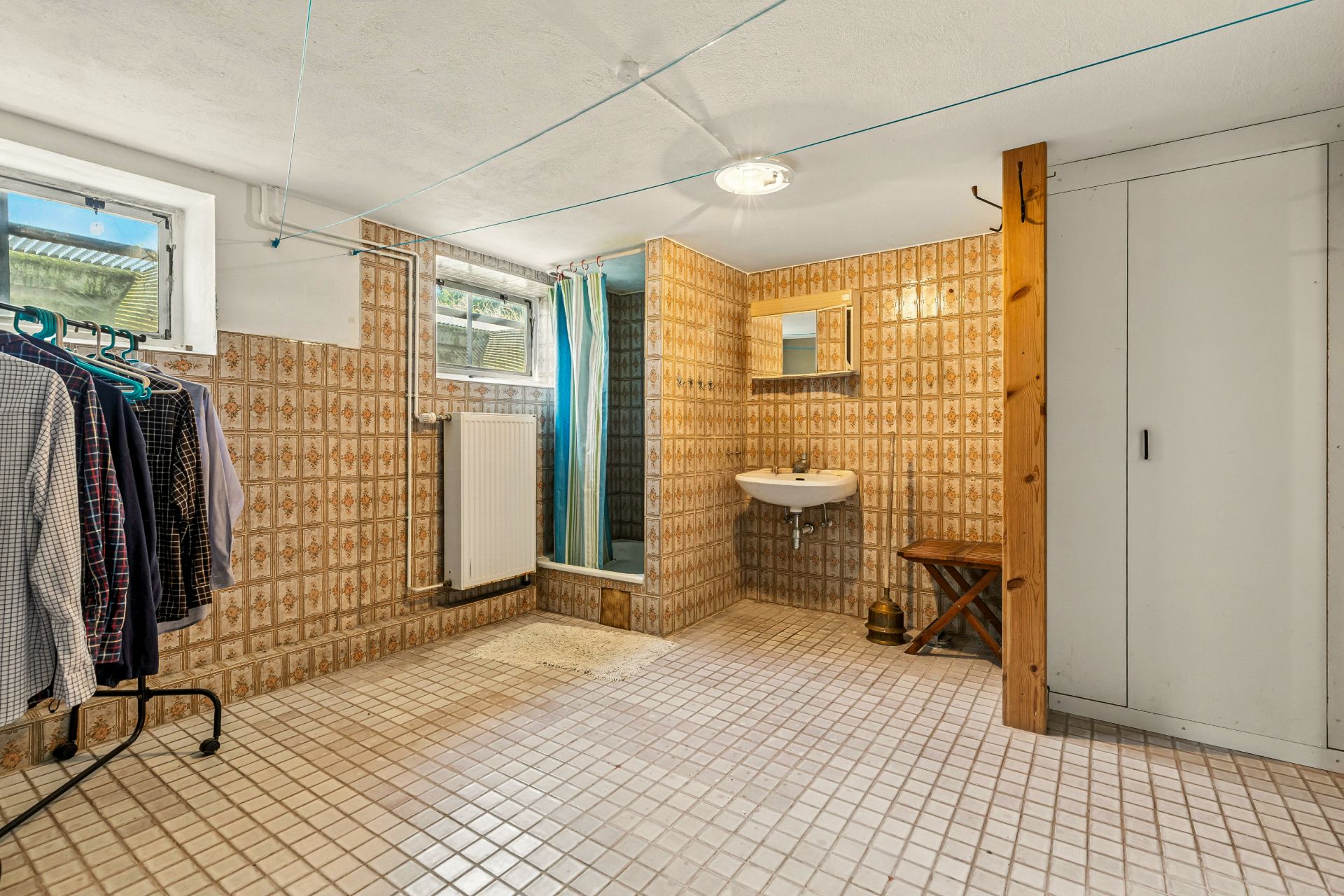 Kellerraum mit Dusche, Waschbecken, Kleiderschrank und Kleiderständer, helle Fliesen.