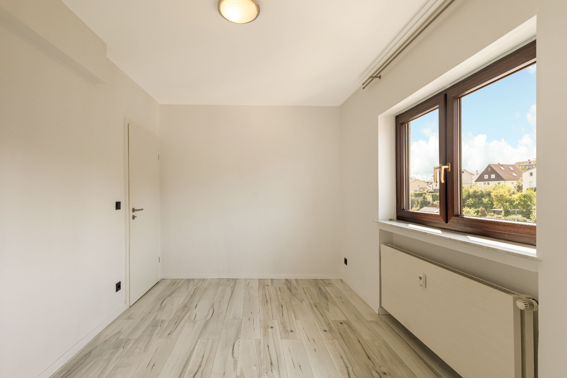 Helles, leeres Zimmer mit Holzfußboden und großem Fenster, durch das Häuser zu sehen sind.
