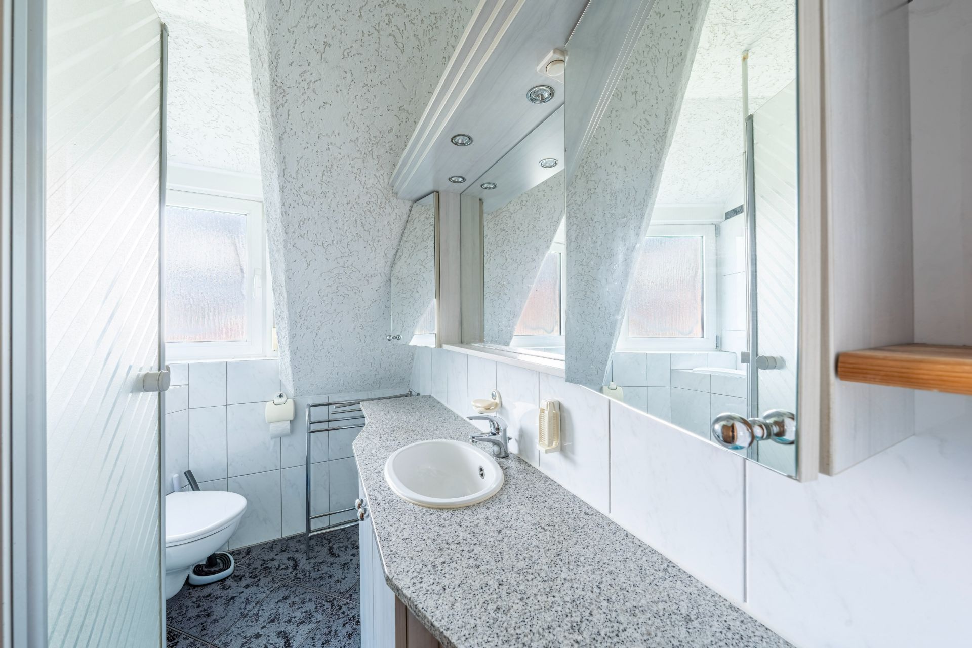 Helles Badezimmer mit Waschbecken, Spiegel, Toilette und zwei Fenstern unter einem schrägen Dach.