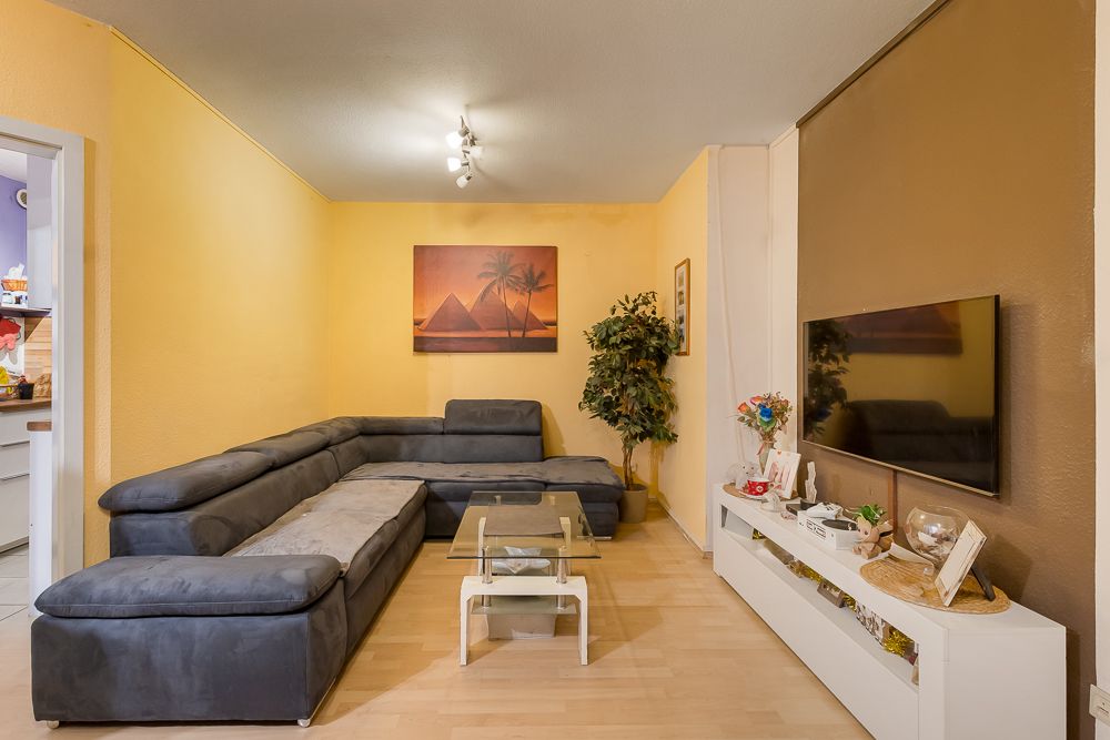 Wohnzimmer mit grauem Ecksofa, Glastisch, TV, gelben Wänden und Wandbild.