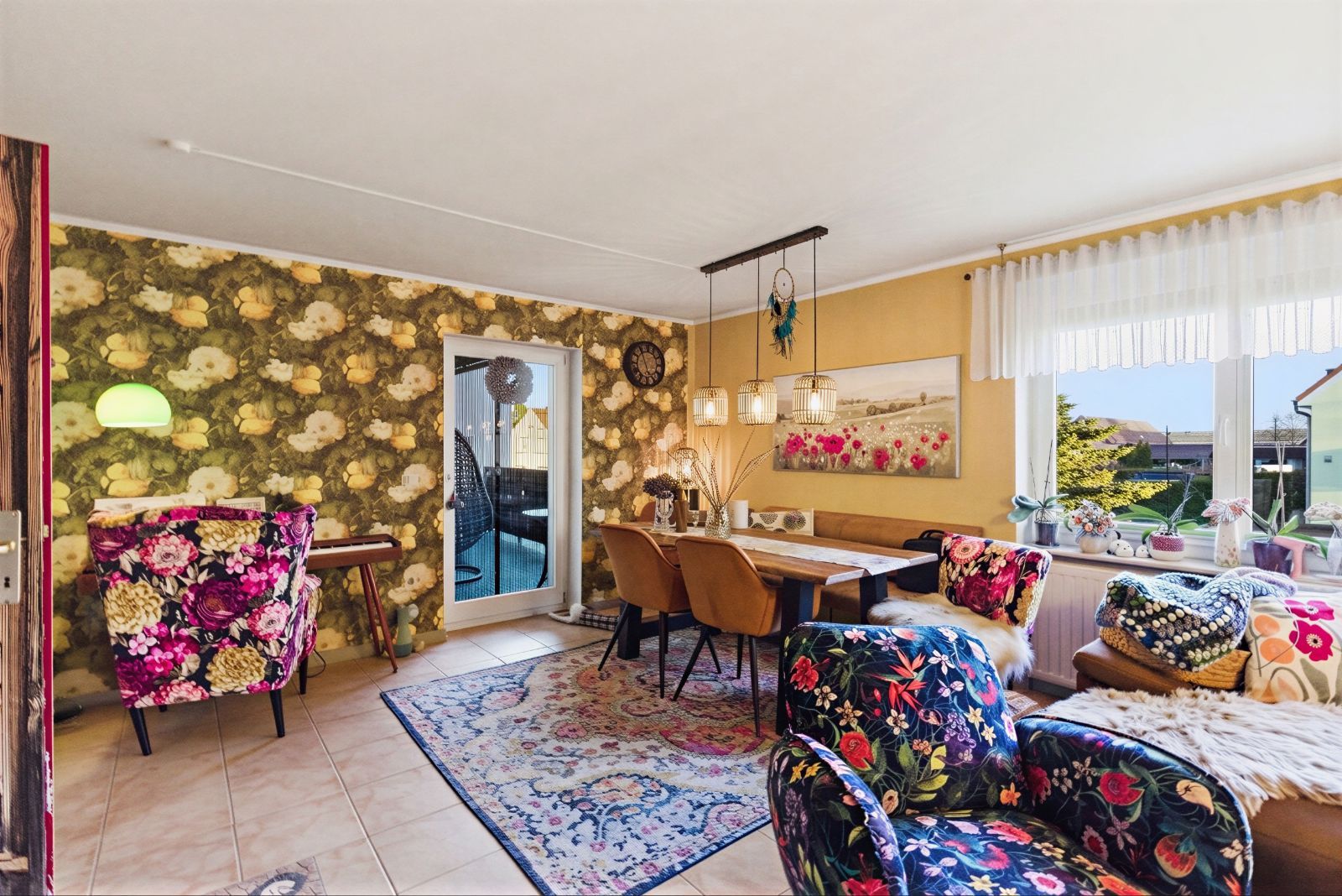 Wohnzimmer mit floral gemusterten Sesseln, Esstisch, Teppich und großem Fenster mit Blick nach draußen.