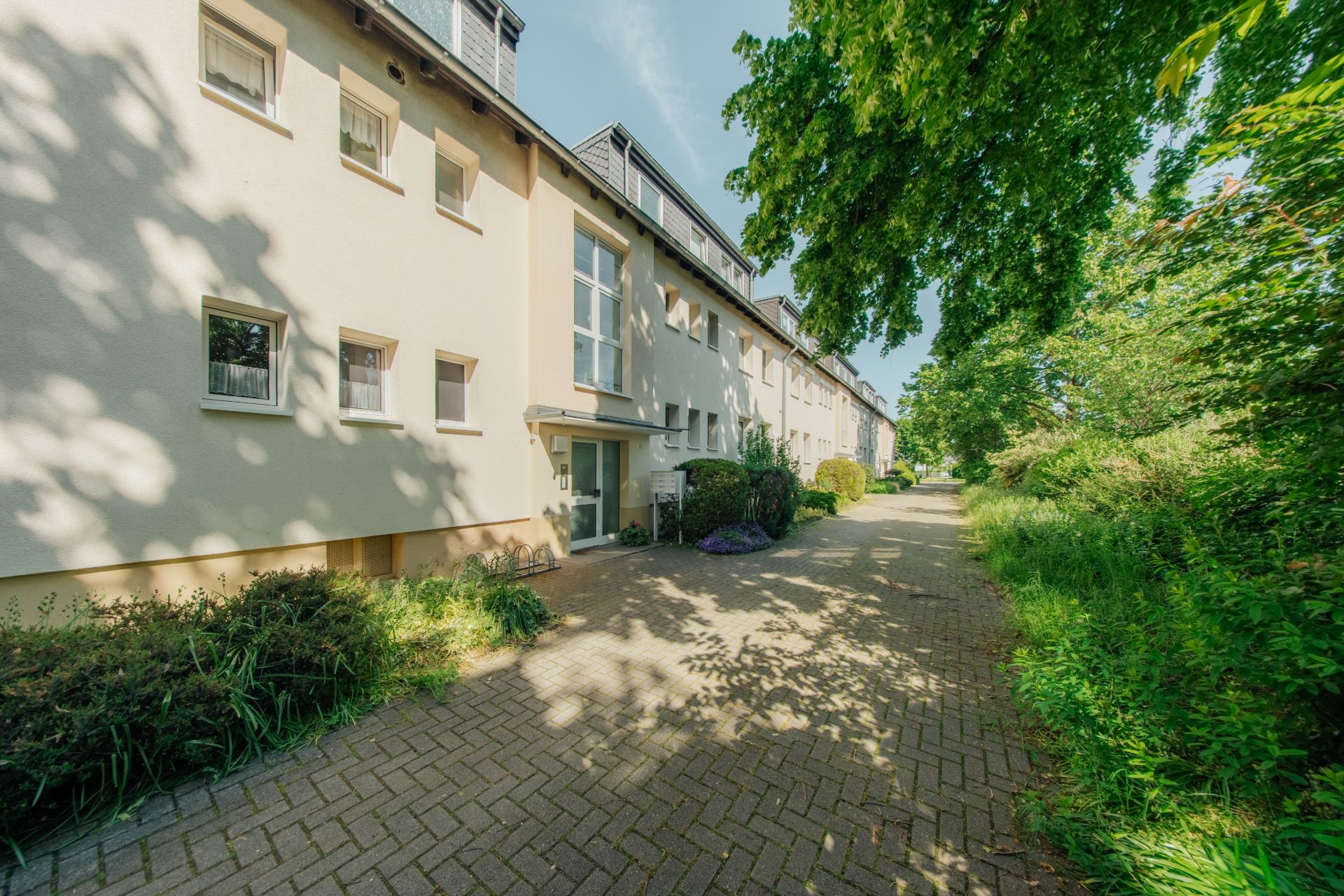 Mehrfamilienhaus mit hellem Fassadenanstrich und gepflastertem Weg, umgeben von Bäumen und Sträuchern.