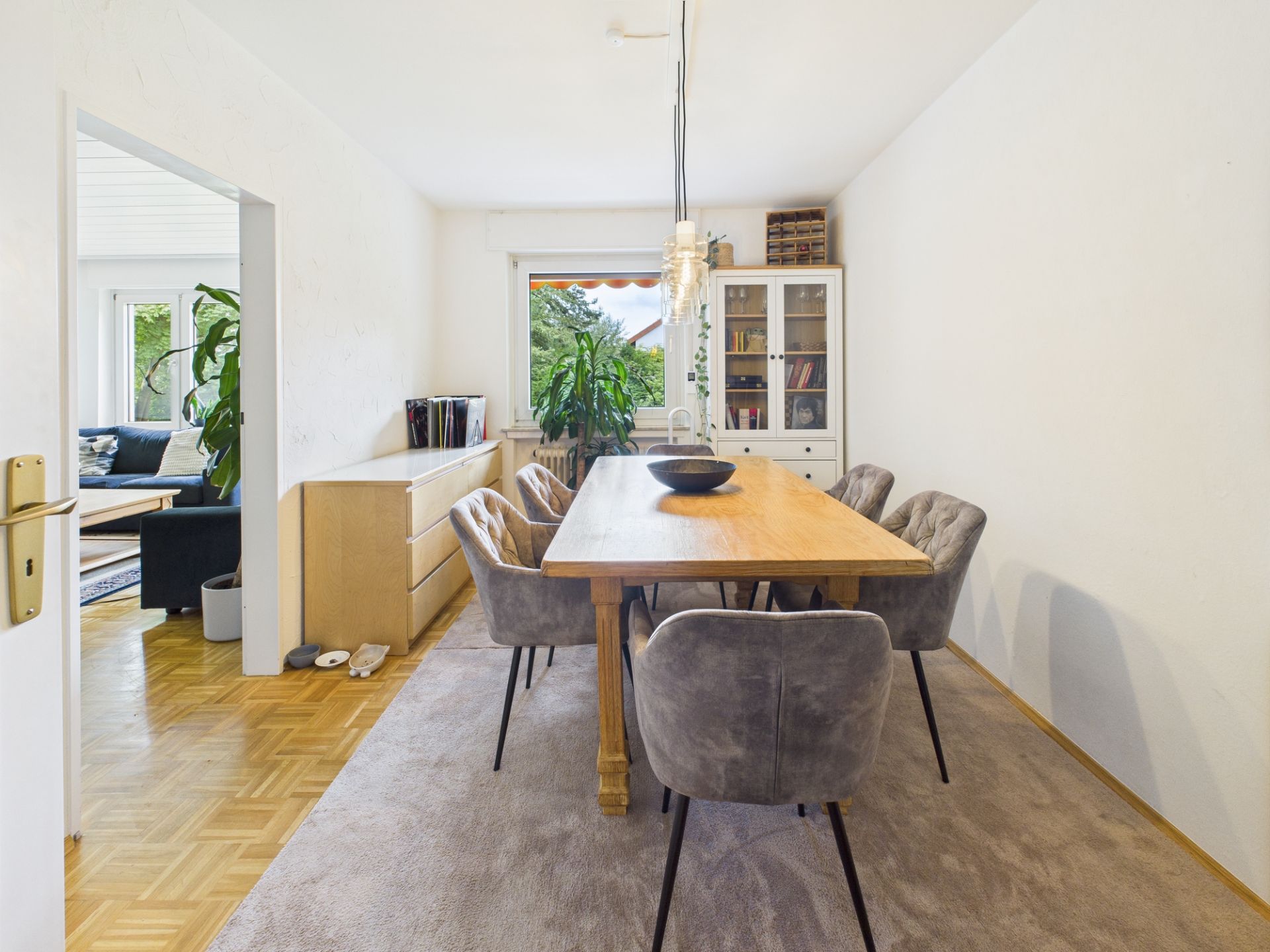 Esszimmer mit Holztisch, sechs Stühlen, Kommode, Vitrine und Fenster mit Pflanzen.