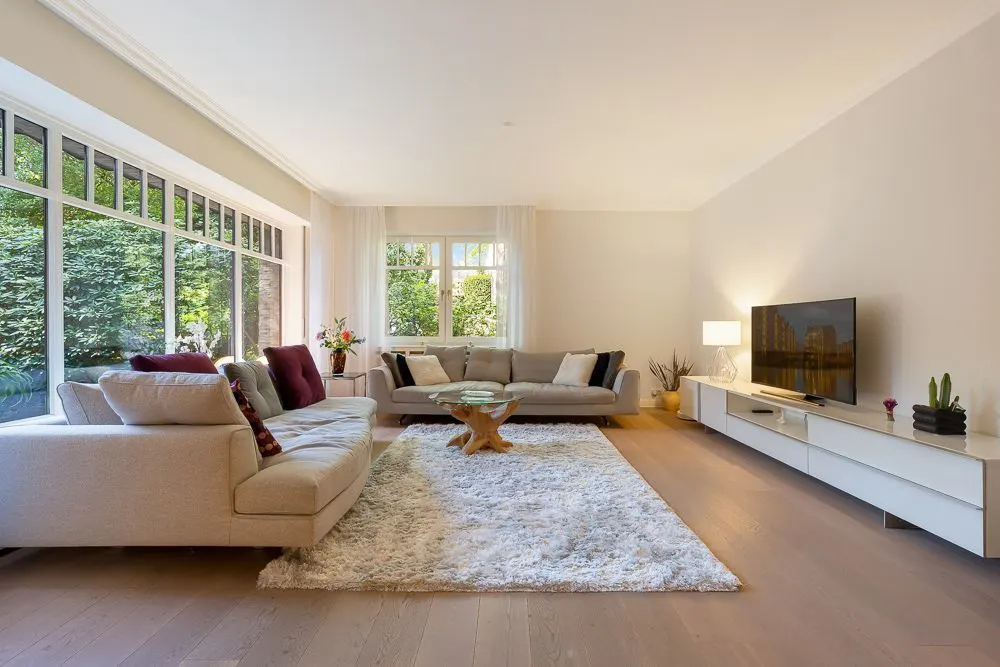 Helles Wohnzimmer mit großen Sofas, Teppich, Couchtisch und Fernseher vor großen Fenstern.