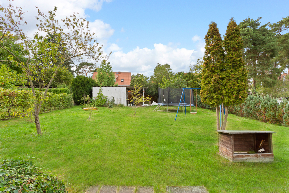 Grüner Garten mit Bäumen, Schaukel, Gartentisch, Trampolin und kleinem Holzhaus.
