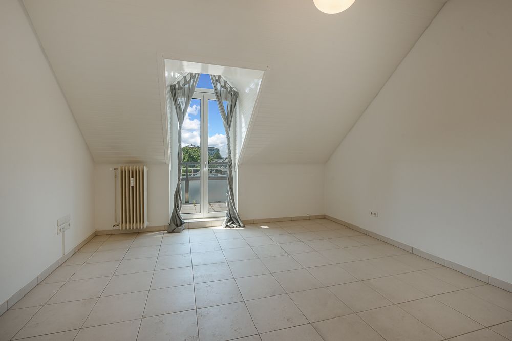Heller leerer Raum mit Fliesenboden, Dachschräge und Fenster mit Vorhängen und Blick ins Freie.