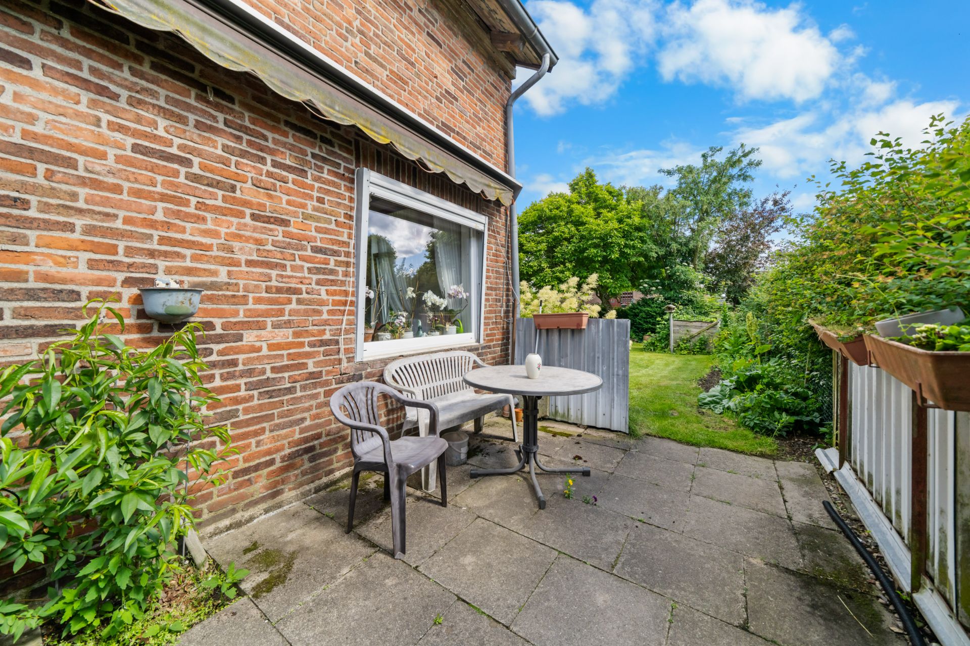 Kleine Terrasse mit Tisch und Stühlen vor einem Backsteinhaus, Blick auf Garten und Zaun.