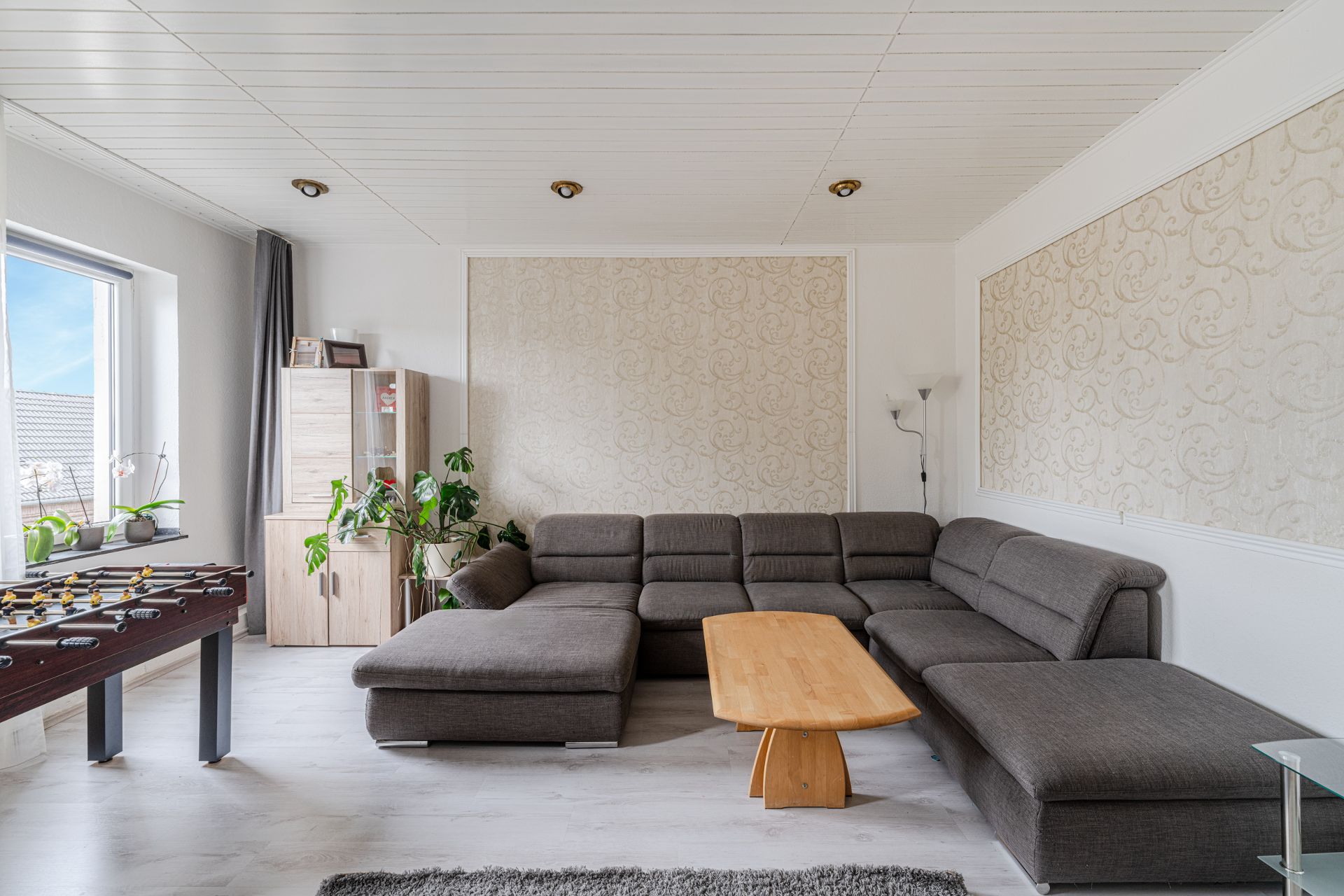 Helles Wohnzimmer mit grauer Eckcouch, Holztisch, Schrank, Pflanzen und Tischkicker am Fenster.