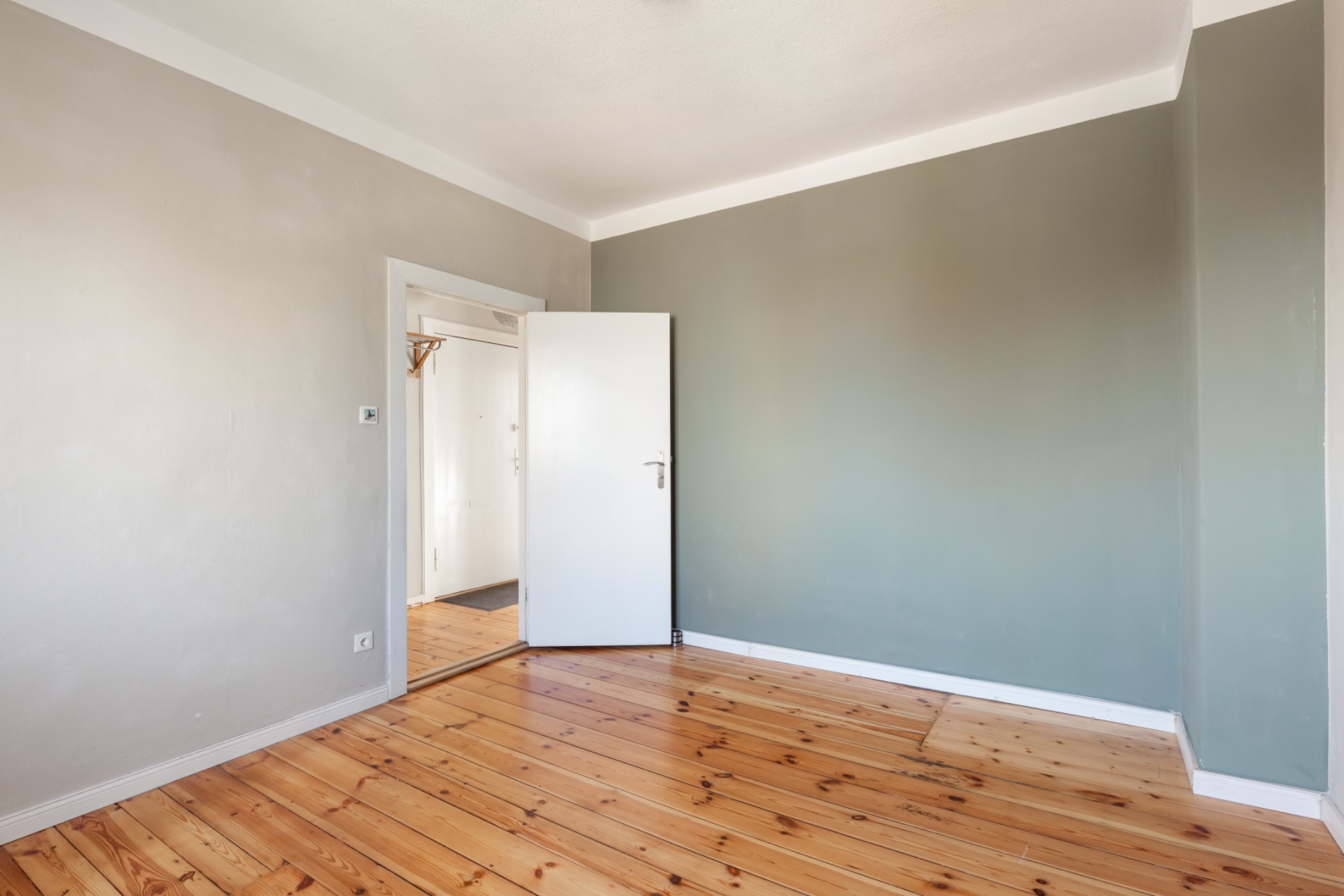 Leerer Raum mit Holzboden, hellgrüner Wand und geöffneter weißer Tür.