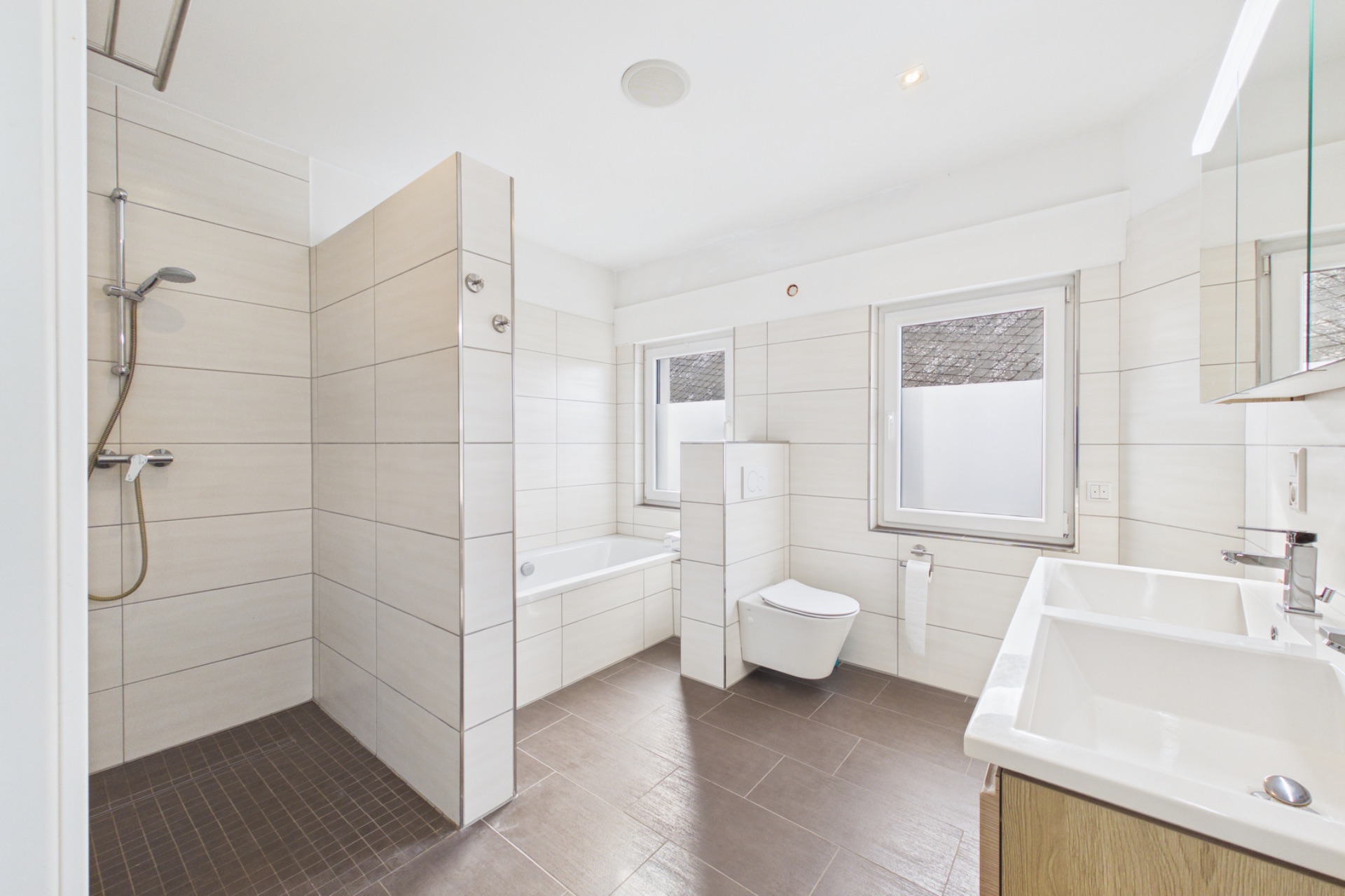 Modernes Badezimmer mit Dusche, Badewanne, WC, zwei Fenstern und Doppelwaschbecken.