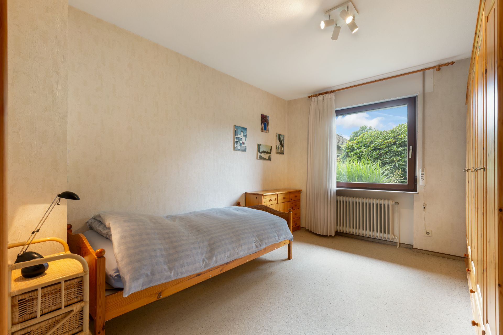Ein einfach eingerichtetes Schlafzimmer mit Einzelbett, Holzschrank, Schreibtisch und Fenster mit Blick ins Grüne.