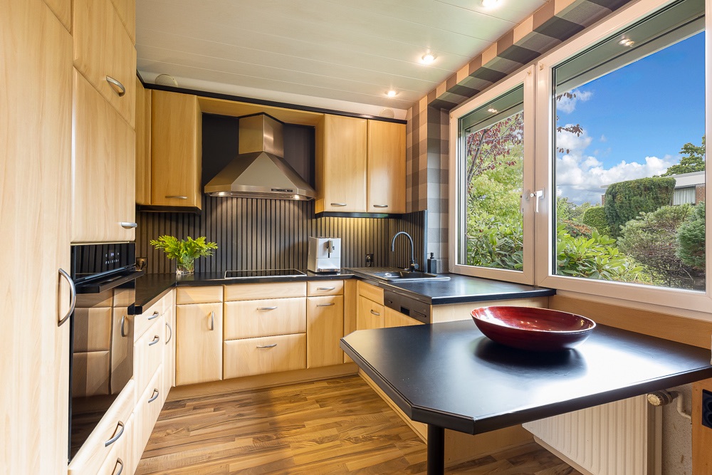 Moderne Küche mit Holzschränken, großem Fenster und schwarzer Arbeitsfläche mit roter Schale.