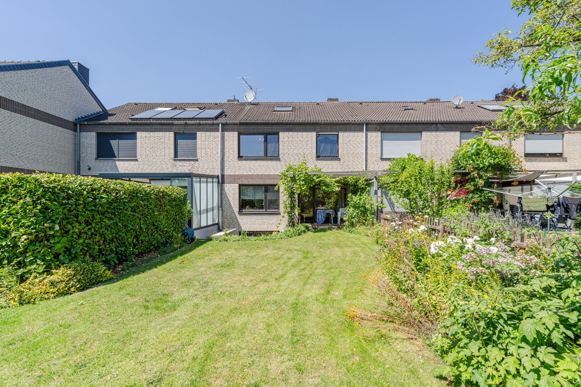 Reihenhaus mit Garten, Rasenfläche, Hecke und Blumenbeeten bei sonnigem Wetter.