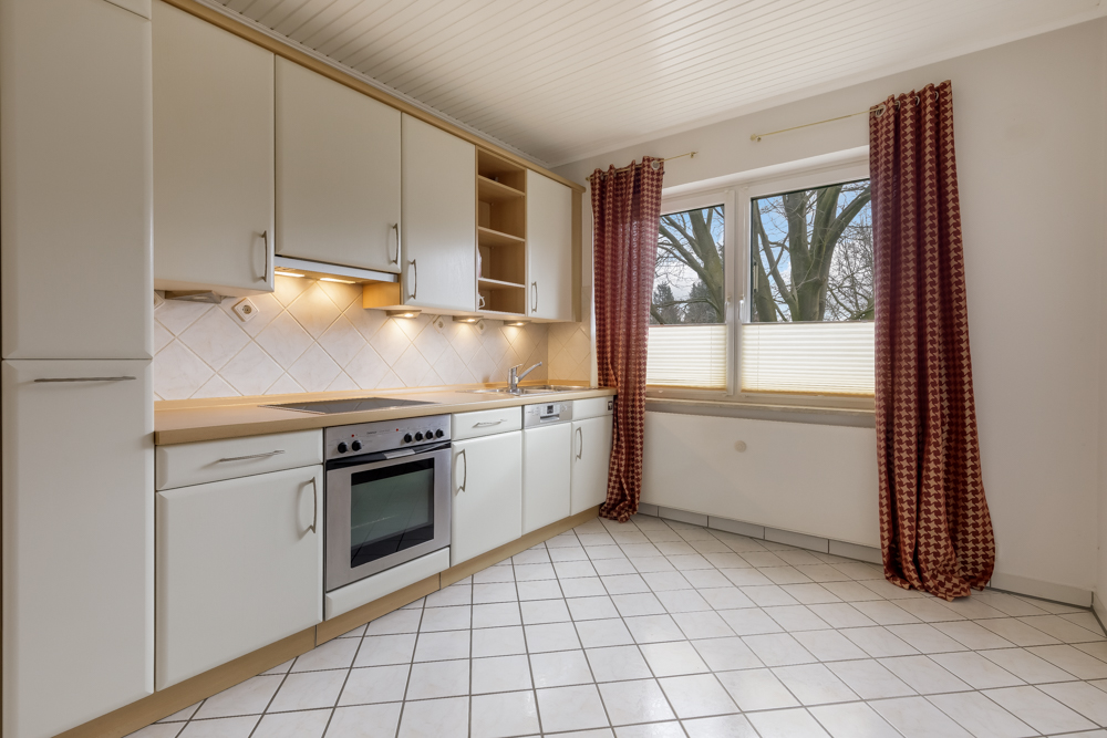 Helle Einbauküche mit weißen Schränken, Herd, Spüle und großem Fenster mit roten Vorhängen.