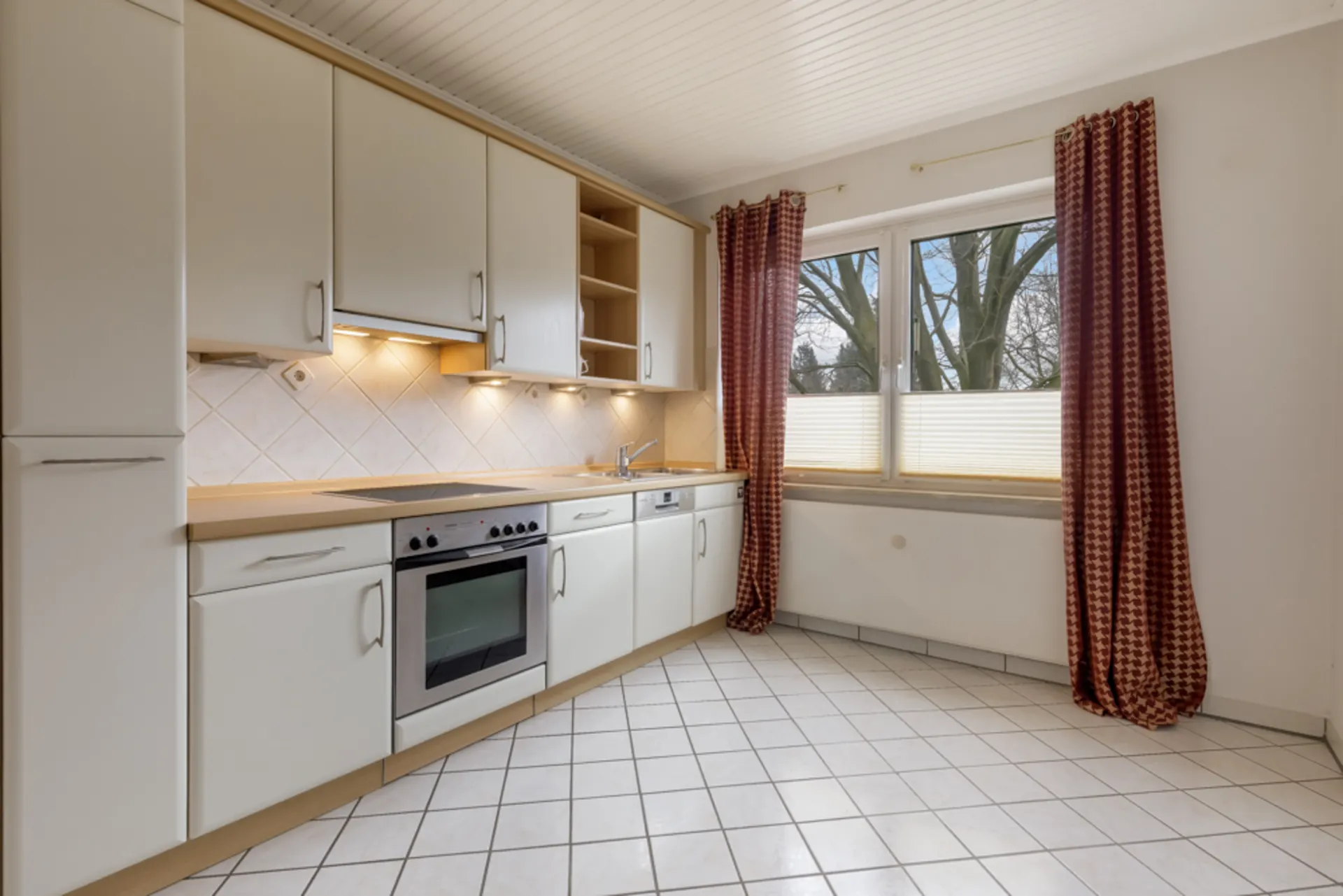 Helle Einbauküche mit weißen Schränken, Herd, Spüle und großem Fenster mit roten Vorhängen.